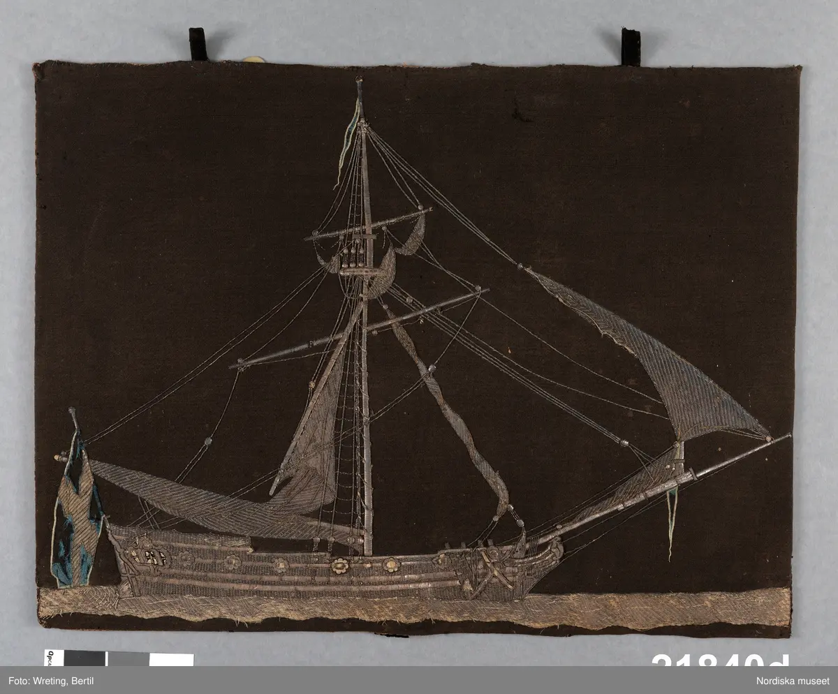 Prydnad till likkista.  Broderad  och monterad på styvt underlag för att hängas på sidan av kistan, med en pendang litt e på andra sidan samt ett mindre med dödskalle och årtalet 1760 , litt c. .
 Bottentyg av mörkbrunt, ursprungligen antagligen svart  tuskaftat ylletyg. Stort fartyg   broderat med silvertråd i olika metallsömstekniker   på ett underlag av pergament för att få reliefverkan  Fartyget som har halade segel är enmastat med gaffelsegel och råsegel  samt  flera försegel på ett långt bogspröt. I aktern den svenska flaggan med applikation av blått sidentyg. Även en toppvimpel med blått siden. Broderiet monterat på en styv pappskiva med baksida av brun linnelärft.  I överkanten två  öglor av svart bomullssammet  för fastsättning på kistan.Troligen är dessa sekundära  Lagningar kring fästet. 
Broderiet är nött och silvertråden svartoxiderad.   Bilderna d och e är identiskt lika men spegelvända.
Förvarades i skepparämbetets i Västerås ämbetskista litt a. 
/Berit Eldvik 2011-08-16