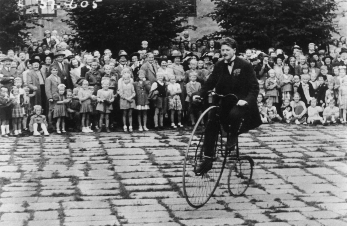 Sykkelkavalkaden på Norsk Folkemuseum i 1951. Publikum på Torvet ser på syklst på gammel sykkel.