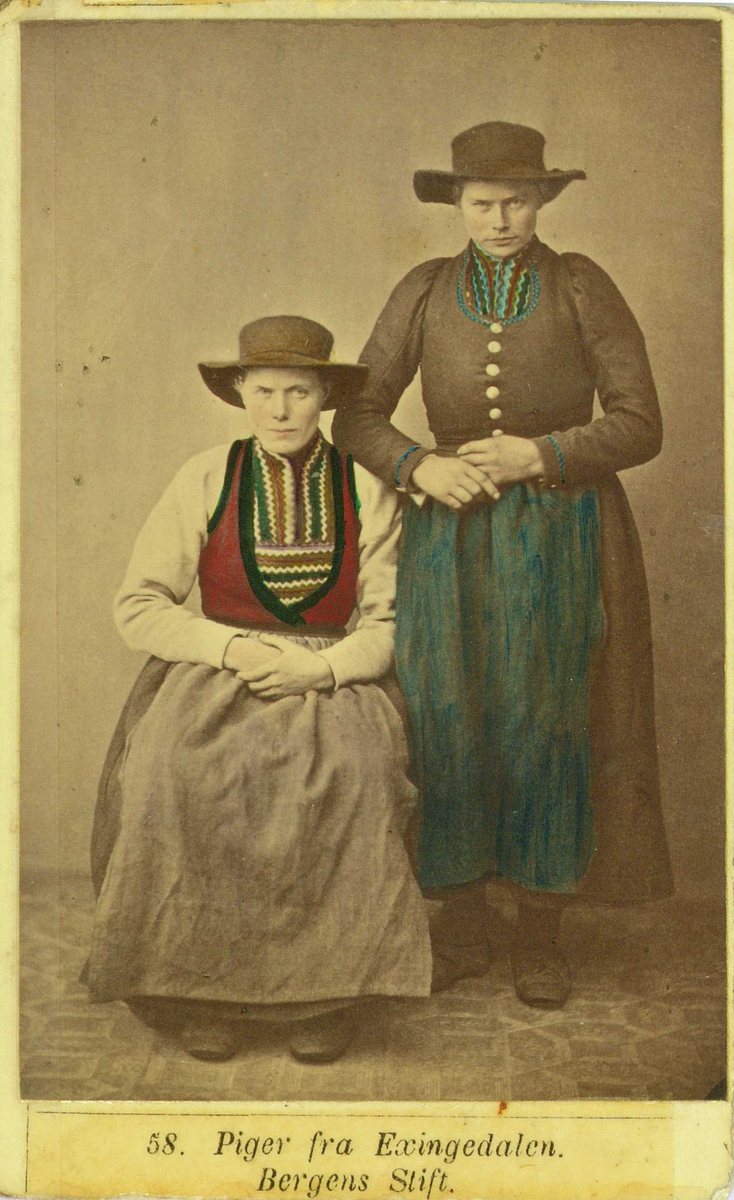 58. Jentedrakter, kvinnedrakter, Eksingedalen, Vaksdal. Portrett av 2 jenter/kvinner med vidbremmede hatter i fotoatelier med nøytral bakgrunn og mønstret gulvteppe. Håndkolorert.
Fra M. Selmers (1819-1900)
folkedraktserie "Norske Nationaldragter", (nr.58).