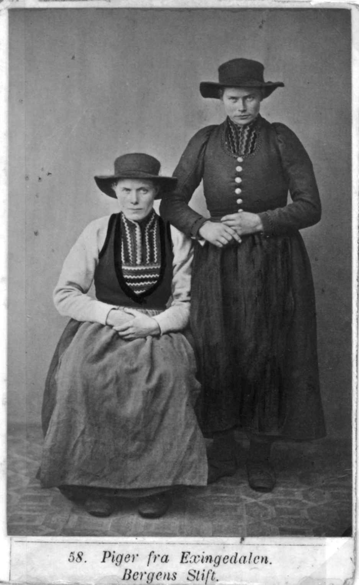 58. Jentedrakter, kvinnedrakter, Eksingedalen, Vaksdal. Portrett av 2 jenter/kvinner med vidbremmede hatter i fotoatelier med nøytral bakgrunn og mønstret gulvteppe. Håndkolorert.
Fra M. Selmers (1819-1900)
folkedraktserie "Norske Nationaldragter", (nr.58).