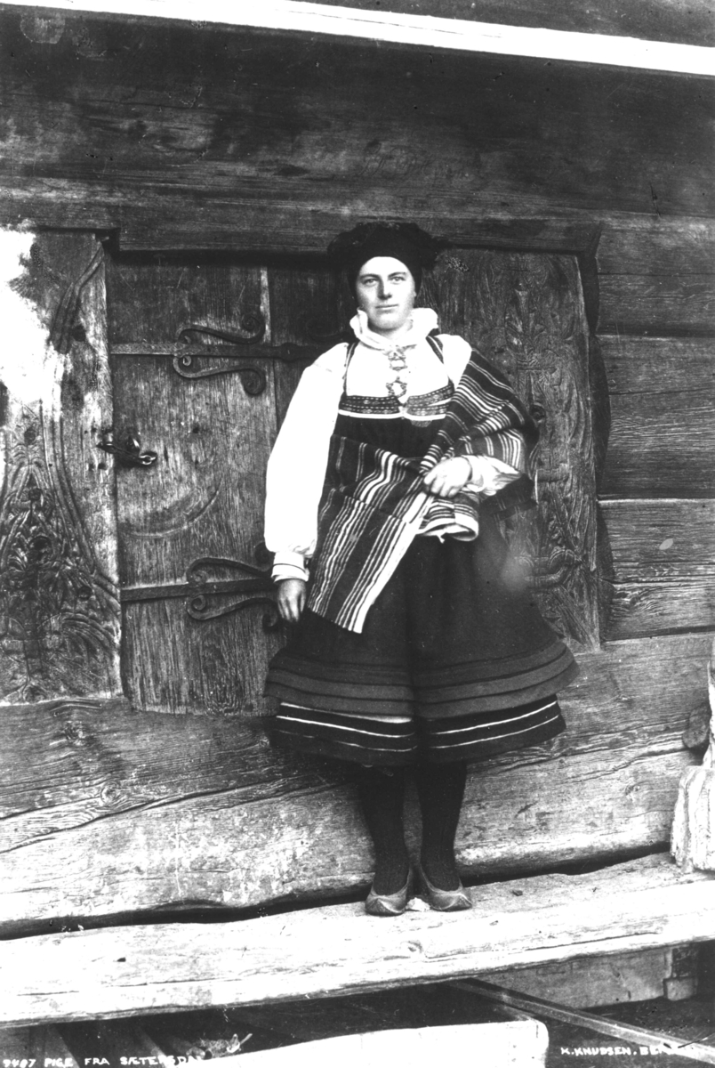 Kvinnedrakt, Setesdal, Aust-Agder. Kvinnen poserer foran loftsdør med beslag.