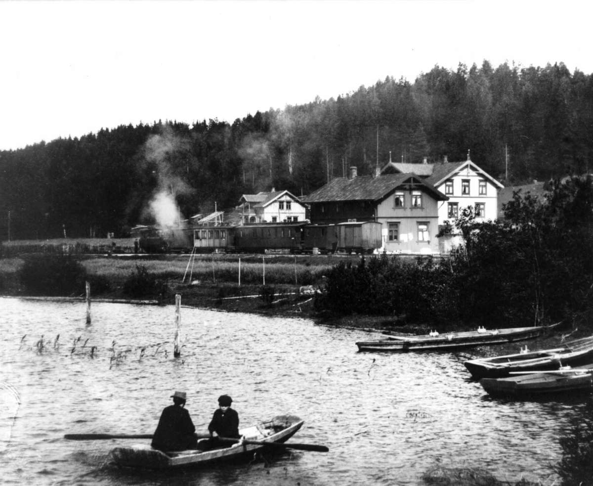 Skullerud, Aurskog-Høland, Akershus 1904. To mennesker i robåt på vann foran jernbanestasjon. Urskog-Hølandsbanen.