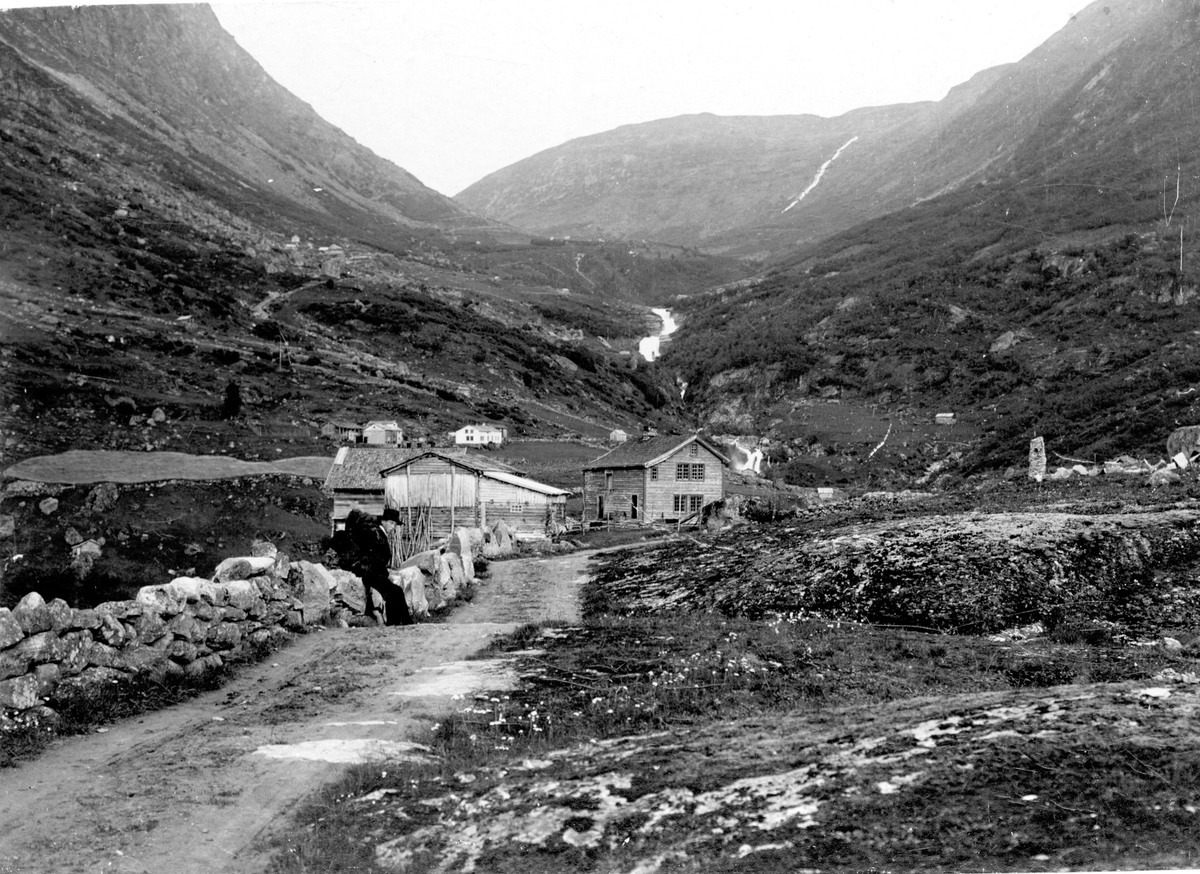 Landskap i Luster, Sogn og Fjordane, på veien gjennom Bergsdalen, mellom Fortun og Turtagrø. Gårdsbebyggelse. Mann sitter på steingjerde i forgrunnen. Gården er en av tre bruk på Berge i Bergsdalen i Luster. I dagligtale kalt Haugen.
Del av gave fra Den norske Turistforening 1972.