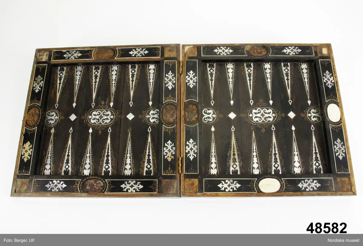 Brädspelslåda för bräde, kvarn och schack, med sju spelbrickor. Svart fanér med inläggningar av pärlemor, elfenben samt graverade förgyllda metallskivor. Blindträ av ek. (Uppgifter från katalogkort). Tillhörande träask för förvaring av spelpjäserna.