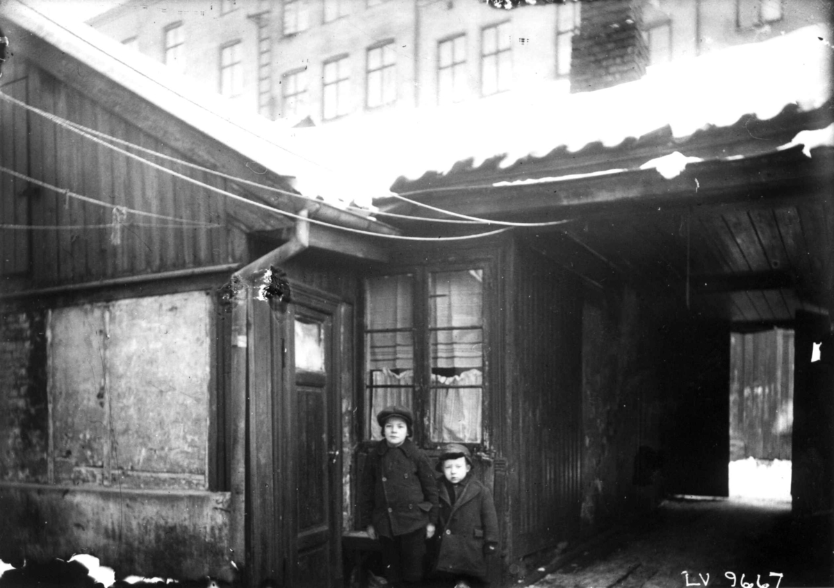 Gårdsrom, bolighus, lav trebebyggelse med høye murgårder i bakgrunnen. Barn poserer foran inngangsdør.
Fra boliginspektør Nanna Brochs boligundersøkelser i Oslo 1920-årene.