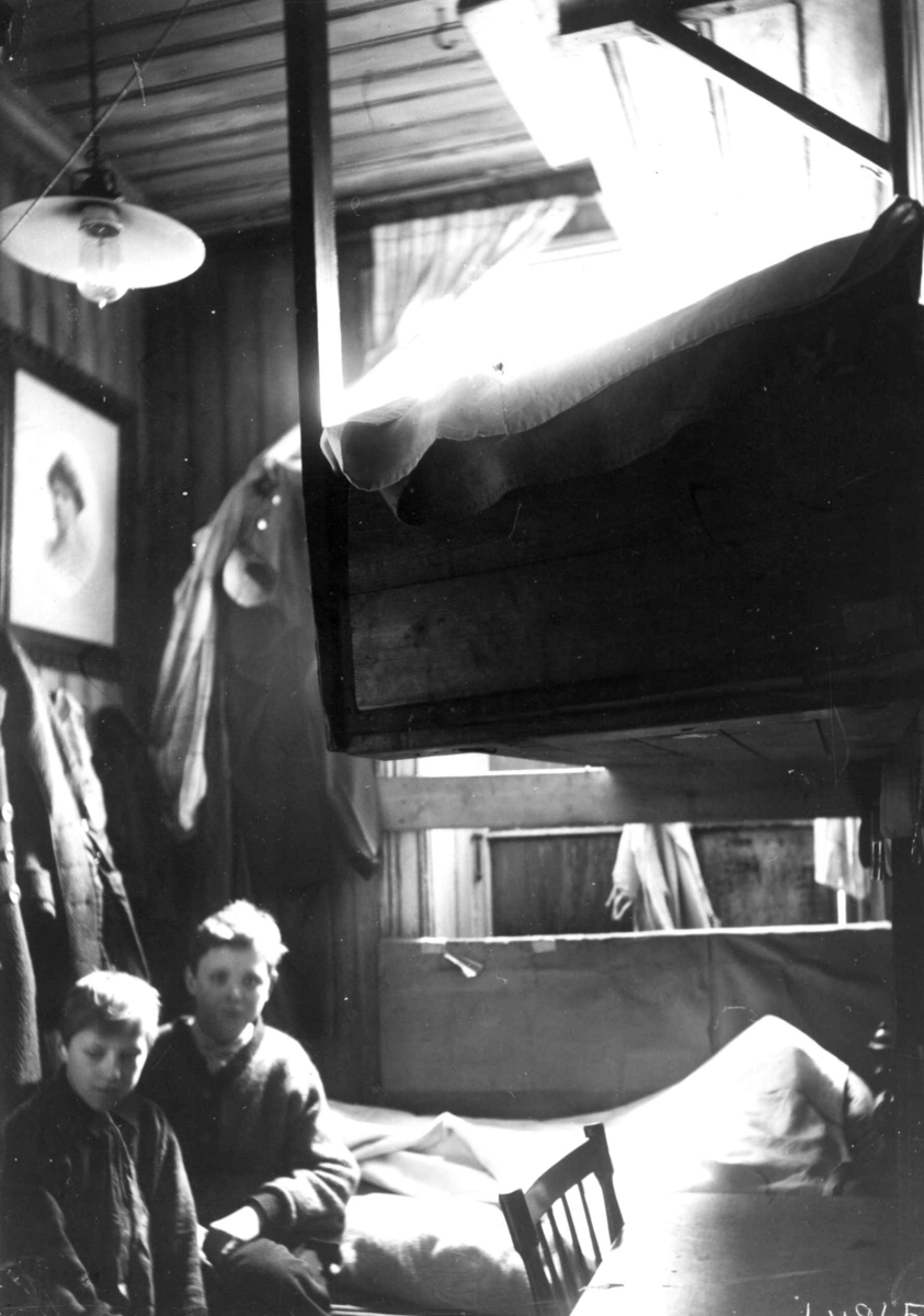 Interiør, Grønlandsstrøket, Oslo. Bebodd bakrom i butikk, 2 gutter sitter mellom seng og kjøkkenkrok, med sengehems (?), i tett møblert rom.
Fra boliginspektør Nanna Brochs boligundersøkelser i Oslo 1920-årene.