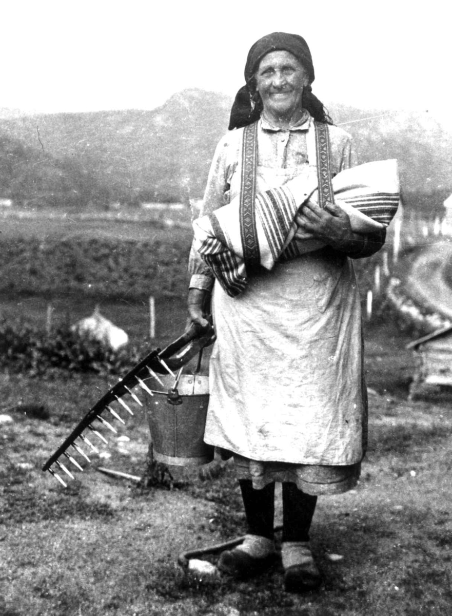 Torbjørg Fidjeland med barn i fatle, i Sirdal kalt linde, og bøtte og rive i den andre hånden. Sirdal, Vest-Agder 1936. Rekonstruksjon.