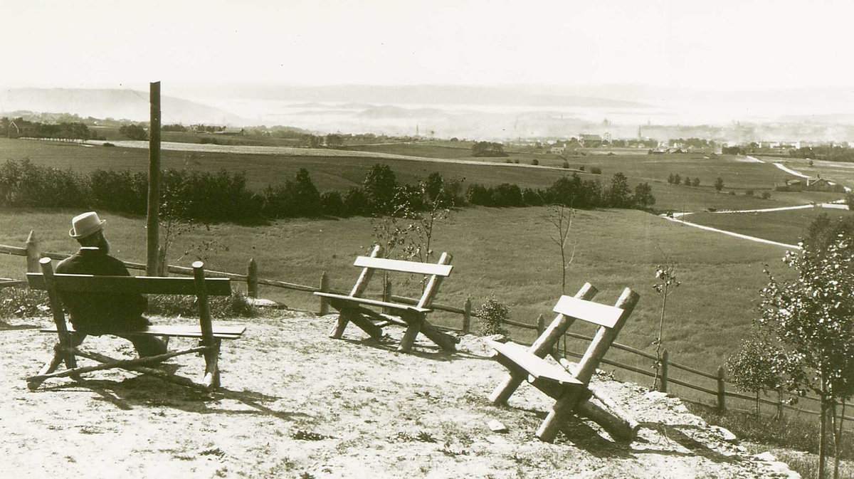 Utsikt over Oslo ant. fra Grefsen. En mann sitter på en benk på et platå i forgrunnen med åpne marker mellom seg og byen i det fjerne. Del av Brødrene Brunskows serie norske landskapsbilder.