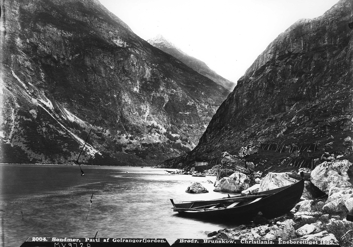 Landskap, del av Geirangerfjorden med robåt trukket opp i fjæresteinene. Kvinne med rive står i h.ytterkant av bildet, nær to rader hesjer i fjellsiden. Sjøbu i bakgrunnen. Del av Den Norske Turistforenings samling landskapsbilder, de fleste tatt av fotografene Brødrene Brunskow.