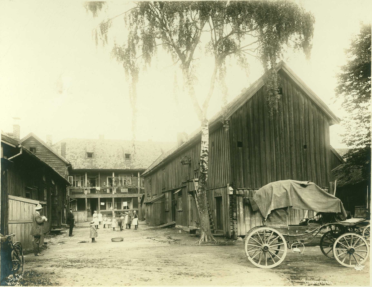 Bygård, Drammen, Buskerud. Trebygninger rundt gårdsrom med barn og voksne. Uthus og bolig med svalgang. Hestekjøretøy plassert i forgrunnen.