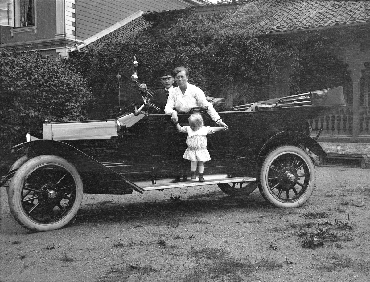 En mann og en kvinne sitter i en åpen bil, kvinnen holder et barn i armene. Robsahm og Lund.