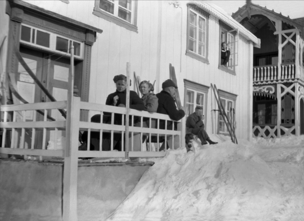 En gruppe mennesker sitter å soler seg utenfor et trehus. Robsahm og Lund.