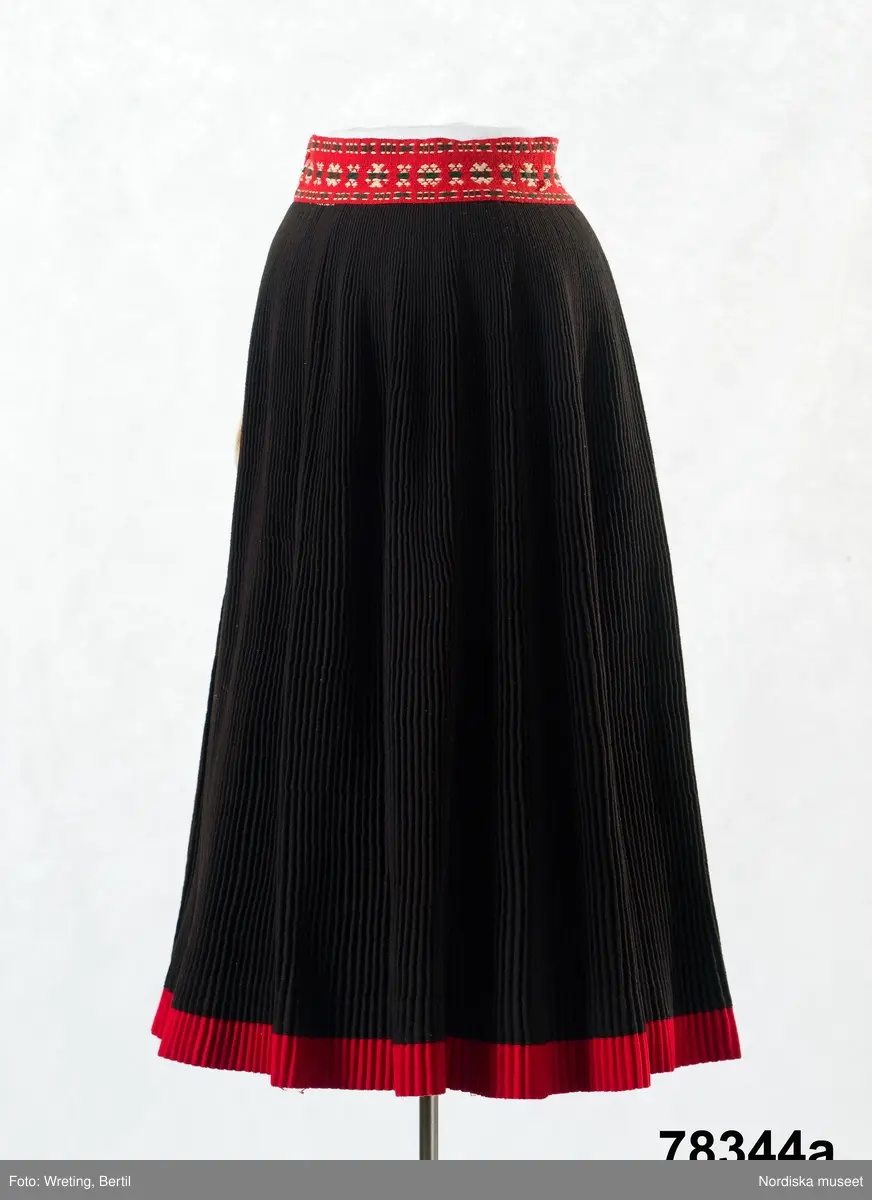 Huvudliggaren:
"Kvinnodräkt fr. Delsbo sn, Norra Helsinglands fgd, Helsingland.
a. "Ryntjeskjortel" af svart kläde med röd kant och bandväfd "kval" (linning). Kjolen "lurad"(goffrerad), skall förvaras lurad att ej vecken gå ut. 3 par fruntimmershänder hjälpas åt att "lura" den efter hvarje begagnande.
b. livstycke, rött med gröna ränder.
c. "Ryntjessärk" (rynksärk) af hvitt bomullstyg med "krus" (handbroderi) kring hals och händer.Begagnas sommartiden
d. "Bumsförkläde" af ylle, blårandigt med bandväf,"kval" i rödt och grönt.
e. "Förklesband" rödt och grönt ylle med "doppar" (tofsar) i ändarna. Lindas 1 à 2 hvarf kring lifvet och bindes framtill med stora nedhängande "locor" och ändar. Längd 262 cm, bredd 3,4 cm.
f. Kjolsäck med mässingslås och hake samt tennbroderi. Häktas under förklesbandet och hänger delvis under "försklet". Begagnas vanligen till förvaring af kyrkkryddor m.fl. kyrk-ellerkalassaker.
g. Halfhandskar af svart kläde med silkesbroderi. Begagnas sommartiden.
h. "Påshufva" hvardagsmössa af brokigt kattun. Begagnas med i nacken utslaget hår med några hårtestar hängande fritt framför öronen.
Till dräkten användes röda strumpor samt låga skor med höga klackar. Äfven kan användas silfersmycken kring halsen eller å  bröstet. En á två brokiga silkesdukar brukades omkring halsen nedstoppade under lifstycket, så att kruset kring halsen  på "ryntjessärken" sammanfästadt med ett löfspänne samt en smalare kant af hvarje sidenduk, blevo synliga. Vid handlederna sammanfästes ryntjessärken med röda sidenband eller länkknappar.

a. KJOL 
längd: 87 cm
midjeband bredd: 7 cm
av svart halvylle med svart linvarp och inslag av svart 1-tr. ullgarn, de 2 yttersta stadtrådarna är av oblekt lingarn. 4 goffrerade våder samt en slät framvåd som skarvats under förklädet med oblekt bomullslärft. 5 cm bred list av rött kläde i nederkanten runtom. Brett midjeband mönstervävt i rött och grönt ullgarn och vitt bomullsgarn. Knäppt vid framvådens vänstra sida med 2 hakar och hyskor av mässing.
b. LIVSTYCKE
längd: 39 cm
av halvylle i varptäckande kypert, varp av vitt lingarn, inslag av svart och grönt ullgarn med flaminslag av vitt, 2 fram-och sidstycken, 2 bakstycken avsmalnande till skörtveck med tre par tränsade förstärkningar av svart garn. I midjehöjd ett avsytt veck runtom, knäppt kant i kant med 8 par grova hakar och hyskor av mässing. Foder av ofärgad bomullslärft.
BE

c. ÖVERDEL
Längd: 39 cm. Längd: 58 cm - ärmlängd. Vidd: 56 cm - ärmvidd. Vidd: 114 cm. Höjd: 1,7 cm - kraghöjd. Material: Textil, bomull. Textil, linne - ärmlinning, krage. Bomullsgarn - broderi. Telnik: Tuskaft. Trådbundet broderi. Broderi, rätlinjig plattsöm. Handsömnad.
Överdel av tuskaftat vitt bomullstyg. Dubbelvikt bålstycke, isydda rektangulära halsspjäll. Täta breda stripade  rynkor mot halskrage i bomull, 1,7 cm hög. Kragen har broderier i rätlinjig plattsöm, vitt bomullsgarn över hela ytan, nuggor i kanten, langetterade bågar  i framkant. 16 cm långt sprund fram, tränsad förstärkning nedtill, 3 cm.  Vidsydd ärm med  infällda ärmspjäll, lagda veck över axeln fram och bak. Täta breda stripade rynkor mot 2,5 cm bred ärmlinning. Ärmlinningen har yttäckande vita broderier i rätlinjig plattsöm, rombiska former. Vita knytband i bomull i ärmlinningar och krage. Överdelen är handsydd.
/Inga-Lill Eliasson 2007-11-26


d. FÖRKLÄDE
längd=vådbredd: 82 cm
bredd: 97,5 cm
av halvylle, varp av svart lingarn, inslag i varptäckande kypert av svart 1-tr. ullgarn med smala ränder istarkt blått och en knappt synlig svart tråd med vita flamgarnsfläckar av bomull. Upptill stripade rynkor där de blå bårderna utnyttjats dekorativt i rynkningen. Mönstervävt midjeband i grönt på rött ullgarn , bandet knäpps med 2 hakar och hyskor i vänster sida.
I nederkantens båda hörn infodringar på insidan av svart bomullslärft, för att tyget inte skall rulla sig.



Berit Eldvik 2004