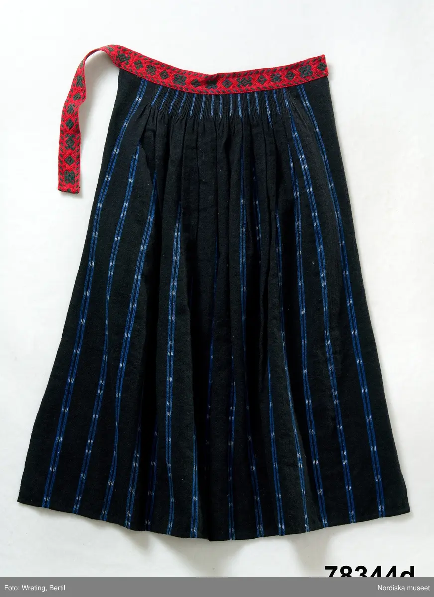 Huvudliggaren:
"Kvinnodräkt fr. Delsbo sn, Norra Helsinglands fgd, Helsingland.
a. "Ryntjeskjortel" af svart kläde med röd kant och bandväfd "kval" (linning). Kjolen "lurad"(goffrerad), skall förvaras lurad att ej vecken gå ut. 3 par fruntimmershänder hjälpas åt att "lura" den efter hvarje begagnande.
b. livstycke, rött med gröna ränder.
c. "Ryntjessärk" (rynksärk) af hvitt bomullstyg med "krus" (handbroderi) kring hals och händer.Begagnas sommartiden
d. "Bumsförkläde" af ylle, blårandigt med bandväf,"kval" i rödt och grönt.
e. "Förklesband" rödt och grönt ylle med "doppar" (tofsar) i ändarna. Lindas 1 à 2 hvarf kring lifvet och bindes framtill med stora nedhängande "locor" och ändar. Längd 262 cm, bredd 3,4 cm.
f. Kjolsäck med mässingslås och hake samt tennbroderi. Häktas under förklesbandet och hänger delvis under "försklet". Begagnas vanligen till förvaring af kyrkkryddor m.fl. kyrk-ellerkalassaker.
g. Halfhandskar af svart kläde med silkesbroderi. Begagnas sommartiden.
h. "Påshufva" hvardagsmössa af brokigt kattun. Begagnas med i nacken utslaget hår med några hårtestar hängande fritt framför öronen.
Till dräkten användes röda strumpor samt låga skor med höga klackar. Äfven kan användas silfersmycken kring halsen eller å  bröstet. En á två brokiga silkesdukar brukades omkring halsen nedstoppade under lifstycket, så att kruset kring halsen  på "ryntjessärken" sammanfästadt med ett löfspänne samt en smalare kant af hvarje sidenduk, blevo synliga. Vid handlederna sammanfästes ryntjessärken med röda sidenband eller länkknappar.

a. KJOL 
längd: 87 cm
midjeband bredd: 7 cm
av svart halvylle med svart linvarp och inslag av svart 1-tr. ullgarn, de 2 yttersta stadtrådarna är av oblekt lingarn. 4 goffrerade våder samt en slät framvåd som skarvats under förklädet med oblekt bomullslärft. 5 cm bred list av rött kläde i nederkanten runtom. Brett midjeband mönstervävt i rött och grönt ullgarn och vitt bomullsgarn. Knäppt vid framvådens vänstra sida med 2 hakar och hyskor av mässing.
b. LIVSTYCKE
längd: 39 cm
av halvylle i varptäckande kypert, varp av vitt lingarn, inslag av svart och grönt ullgarn med flaminslag av vitt, 2 fram-och sidstycken, 2 bakstycken avsmalnande till skörtveck med tre par tränsade förstärkningar av svart garn. I midjehöjd ett avsytt veck runtom, knäppt kant i kant med 8 par grova hakar och hyskor av mässing. Foder av ofärgad bomullslärft.
BE

c. ÖVERDEL
Längd: 39 cm. Längd: 58 cm - ärmlängd. Vidd: 56 cm - ärmvidd. Vidd: 114 cm. Höjd: 1,7 cm - kraghöjd. Material: Textil, bomull. Textil, linne - ärmlinning, krage. Bomullsgarn - broderi. Telnik: Tuskaft. Trådbundet broderi. Broderi, rätlinjig plattsöm. Handsömnad.
Överdel av tuskaftat vitt bomullstyg. Dubbelvikt bålstycke, isydda rektangulära halsspjäll. Täta breda stripade  rynkor mot halskrage i bomull, 1,7 cm hög. Kragen har broderier i rätlinjig plattsöm, vitt bomullsgarn över hela ytan, nuggor i kanten, langetterade bågar  i framkant. 16 cm långt sprund fram, tränsad förstärkning nedtill, 3 cm.  Vidsydd ärm med  infällda ärmspjäll, lagda veck över axeln fram och bak. Täta breda stripade rynkor mot 2,5 cm bred ärmlinning. Ärmlinningen har yttäckande vita broderier i rätlinjig plattsöm, rombiska former. Vita knytband i bomull i ärmlinningar och krage. Överdelen är handsydd.
/Inga-Lill Eliasson 2007-11-26


d. FÖRKLÄDE
längd=vådbredd: 82 cm
bredd: 97,5 cm
av halvylle, varp av svart lingarn, inslag i varptäckande kypert av svart 1-tr. ullgarn med smala ränder istarkt blått och en knappt synlig svart tråd med vita flamgarnsfläckar av bomull. Upptill stripade rynkor där de blå bårderna utnyttjats dekorativt i rynkningen. Mönstervävt midjeband i grönt på rött ullgarn , bandet knäpps med 2 hakar och hyskor i vänster sida.
I nederkantens båda hörn infodringar på insidan av svart bomullslärft, för att tyget inte skall rulla sig.



Berit Eldvik 2004