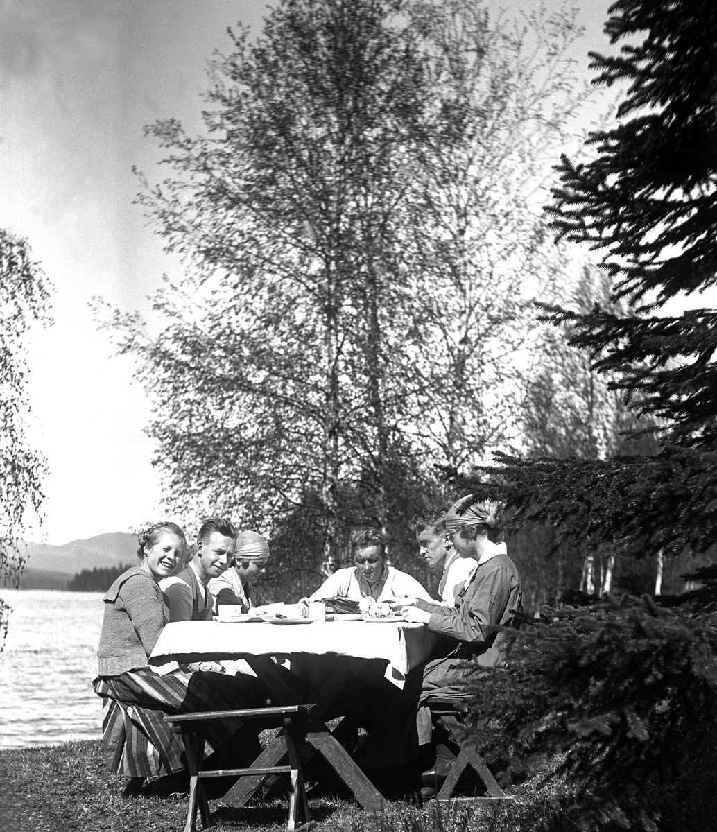 Seks unge mennesker spiser frokost i det fri, antagelig ved Harestuvatnet. Fotografert 1921.