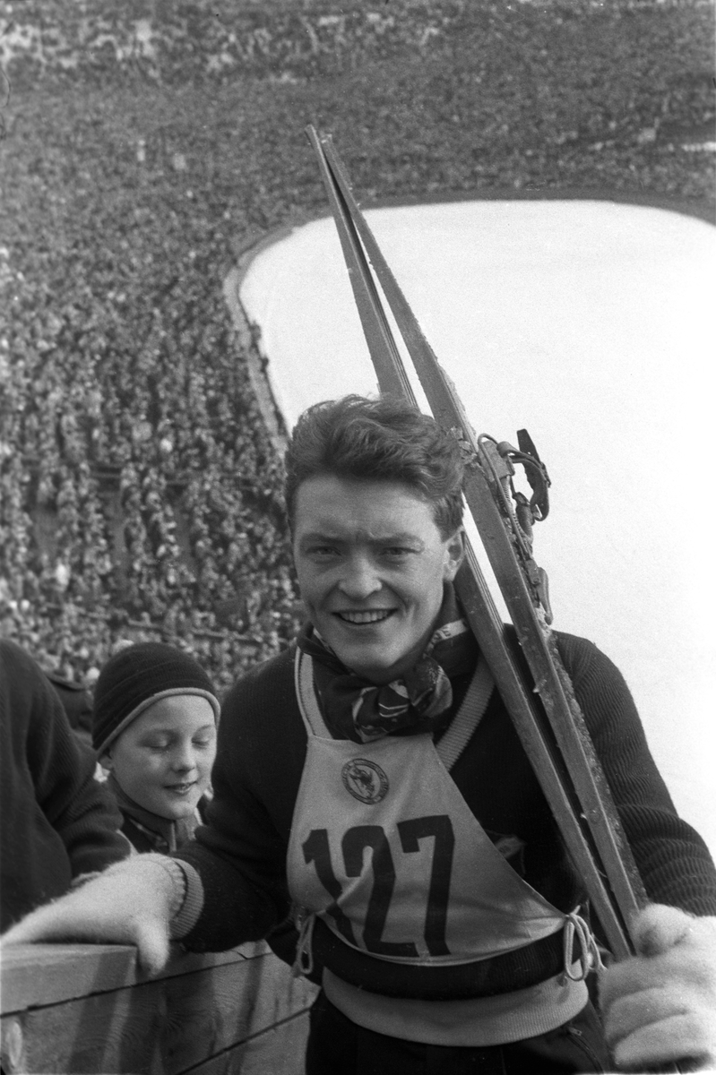 Hopper med startnummer 127 på vei opp i hoppet med skiene på skulderen. Publikum i bakgrunnen. Holmenkollhopp 1953. Fotograf Dagbladet