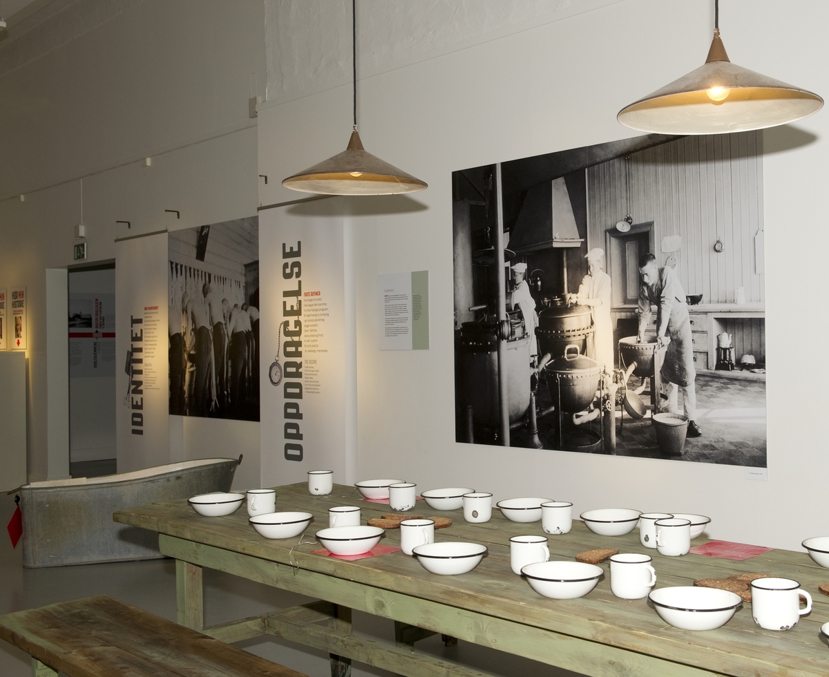 Utstillingsplakater med bilder fra Bastøy skolehjem. Lamper, bord og koppestell er rekvisitter som ble brukt i spillefilmen "Kongen av Bastøy". Fra utstillingen om Bastøy skolehjem på Norsk Folkemuseum. Utstillingen stod i perioden 15.12.2010 til 31.12.2011.