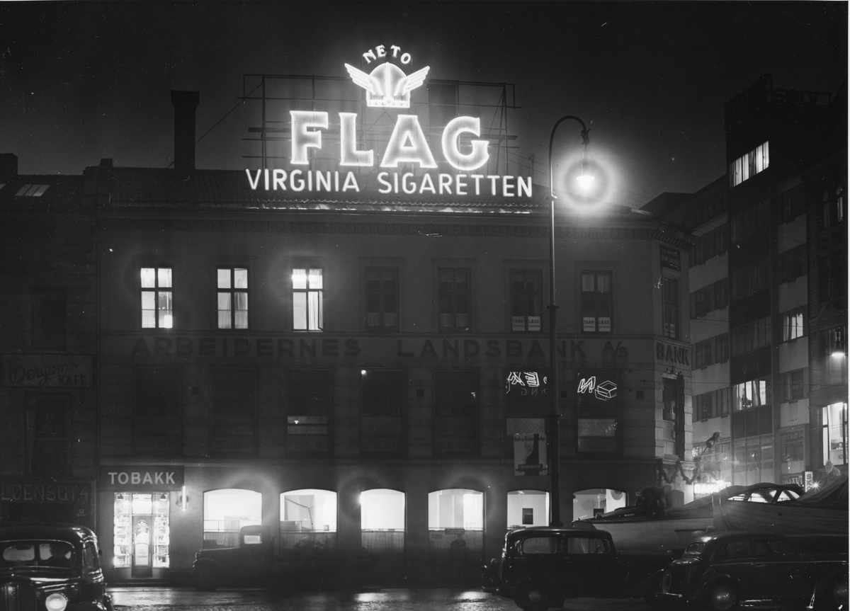 Gatebilde. Hus med Arbeidernes Landsbank A/S, Bergums kafé og en tobakksbutikk. Neonskilt på taket med reklame for NETO Flag sigaretter.