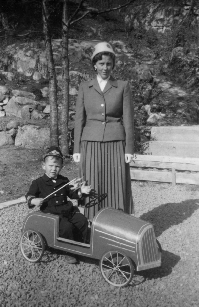 Finn Kreutz med matrosdress fotografert i sin lekebil. Mor, Ingeborg Kreutz, i bakgrunnen. Trolig 17. mai 1958.