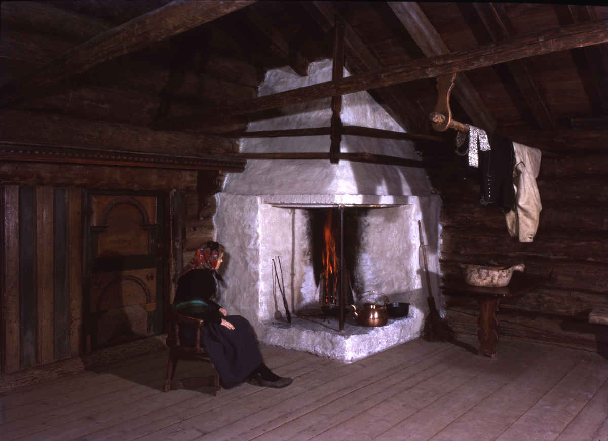 Interiør fra Grøslistua i Numedalstunet i friluftsmuseet på Norsk Folkemuseum. Kvinne sitter ved peis.