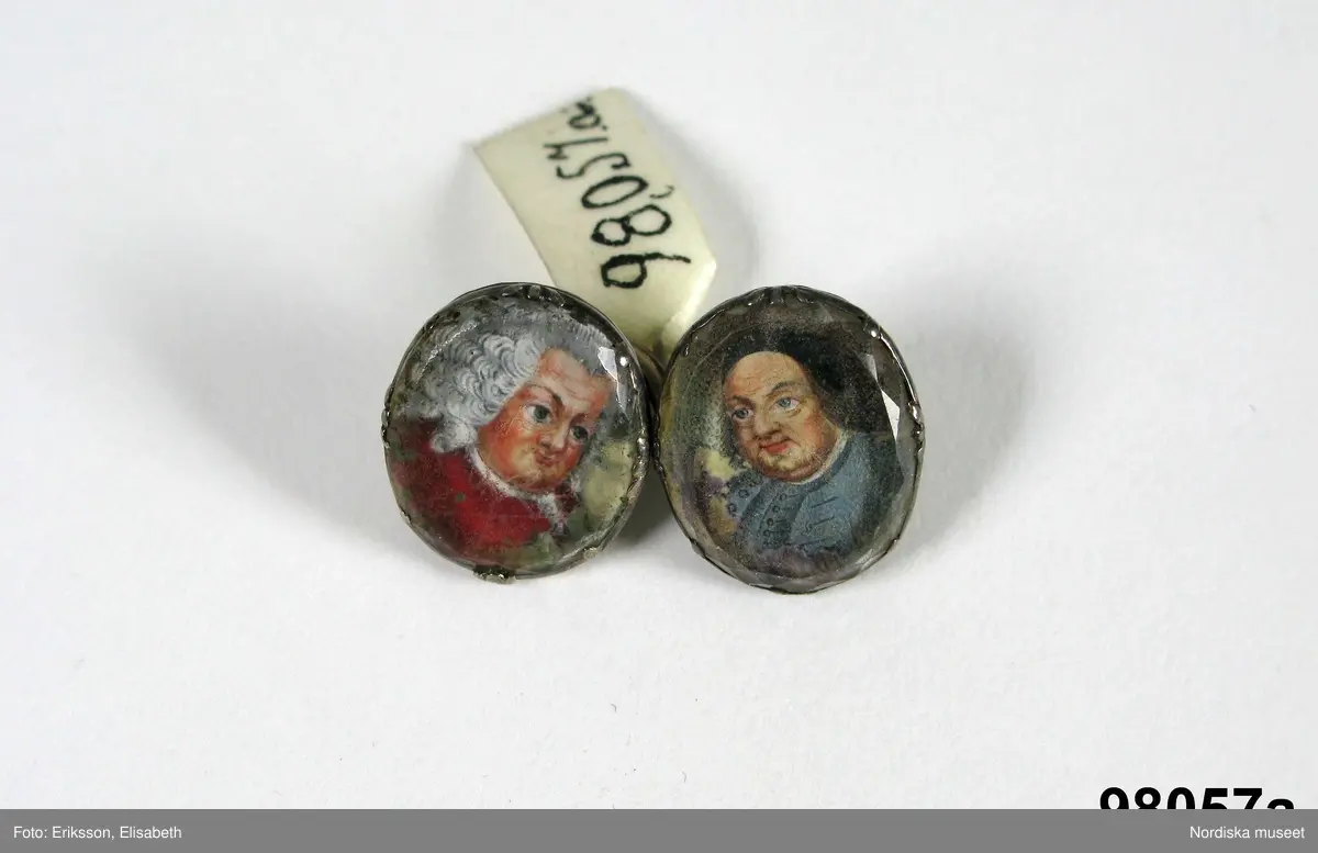 Manschettknappar, två par, miniatyrer. 1700-tals mitt.  Silver, glas.
Saknar stämplar. På varje knapp ett ovalt porträtt i miniatyr i silverinfattning, baksidan räfflad, knapparna förenade två och två, på den ena föreningslänken. ”I H” i genombrutit arbete, den andra föreningslänken senare ersättning. Manschettknapparna hölls i ett rött marokäng (getskinn) etui.
(katalogiseringen gäller a-c)