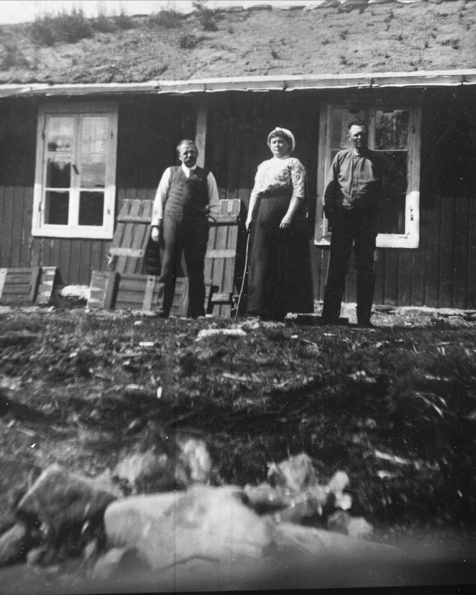 Avfotografering. Menn og en kvinne foran et torvdekket hus.