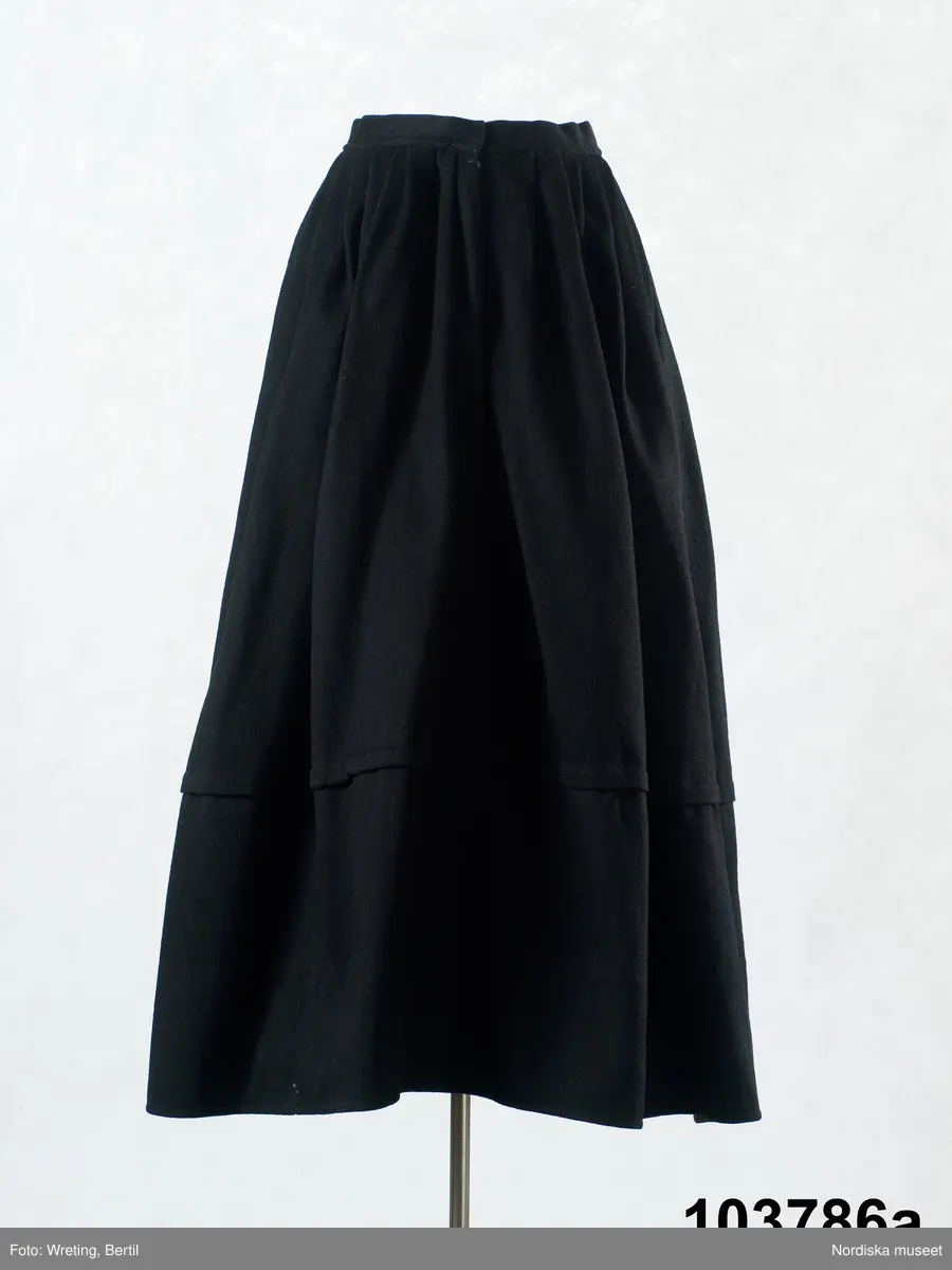 b. Brudtröja av svart vadmal,  Runt alla kanter bård av ytterst brunsvart sammetsband och innanför ett 4 cm brett svart sammetsband  med invävt bladmönster. På ärmarna ett svart slätt sammetsband med pålagd guldspets därefter siden band i rosa och grönt på botten av  svart  sammet och överst i bården en knypplad guldspets. 
c: Förkläde. Material: Textil, bomull. Teknik: Manufakturvävning. Handsömnad. Längd: 77 cm. Bredd: 110 cm. Färg: Vit.
Förkläde i vitt manufakturvävt bomullstyg, ihopsytt i mitten, smalrandigt, stärkt. Rynkningar mot midjelinning i tuskaftat bomullstyg, 1,5 cm bred, fastsydda bomullsband i sidorna. Uppvikt fåll nedtill, 6,5 cm, ovanför fåll, 5 lagda veck med ca 1 cm mellanrum, enkel utdragssöm i kanten av vecken.
/Inga-Lill Eliasson 2008-02-25

e: Krage. Längd: 47,5, cm - totallängd, bröststycke. Bredd: 33 cm - bröststycke. Höjd: 14 cm - kraghöjd, inkl spets. Material: Textil, bomull. Lingarn - spets. Bomullsgarn - broderi. Teknik: Tuskaft. Vitbroderi, Knyppling. Handsömnad.
Krage i tuskaftat vitt bomullstyg. Rektangulär duk, uppklippt halshål med fastsydd krage, 14 cm hög. Kragen har 3,5 cm bred fåll, enkel utdragssöm i kanten. Vitbroderi  i framkant,  plattsöm och kantsydda hål med infällt tyll, två sydda knapphål. Handknypplad spets fastsydd på långsida och korstsidor, 2,5 cm bred. Öppning fram, 24 cm långt sprund. Kragen är handsydd.
Lästips: Svensson Sigfrid, (1935). Skånes Folkdräkter, en dräkthistorisk undersökning 1500 - 1900. Stockholm: Nordiska Museets Handlingar: 3.
/Inga-Lill Eliasson 2008-02-19