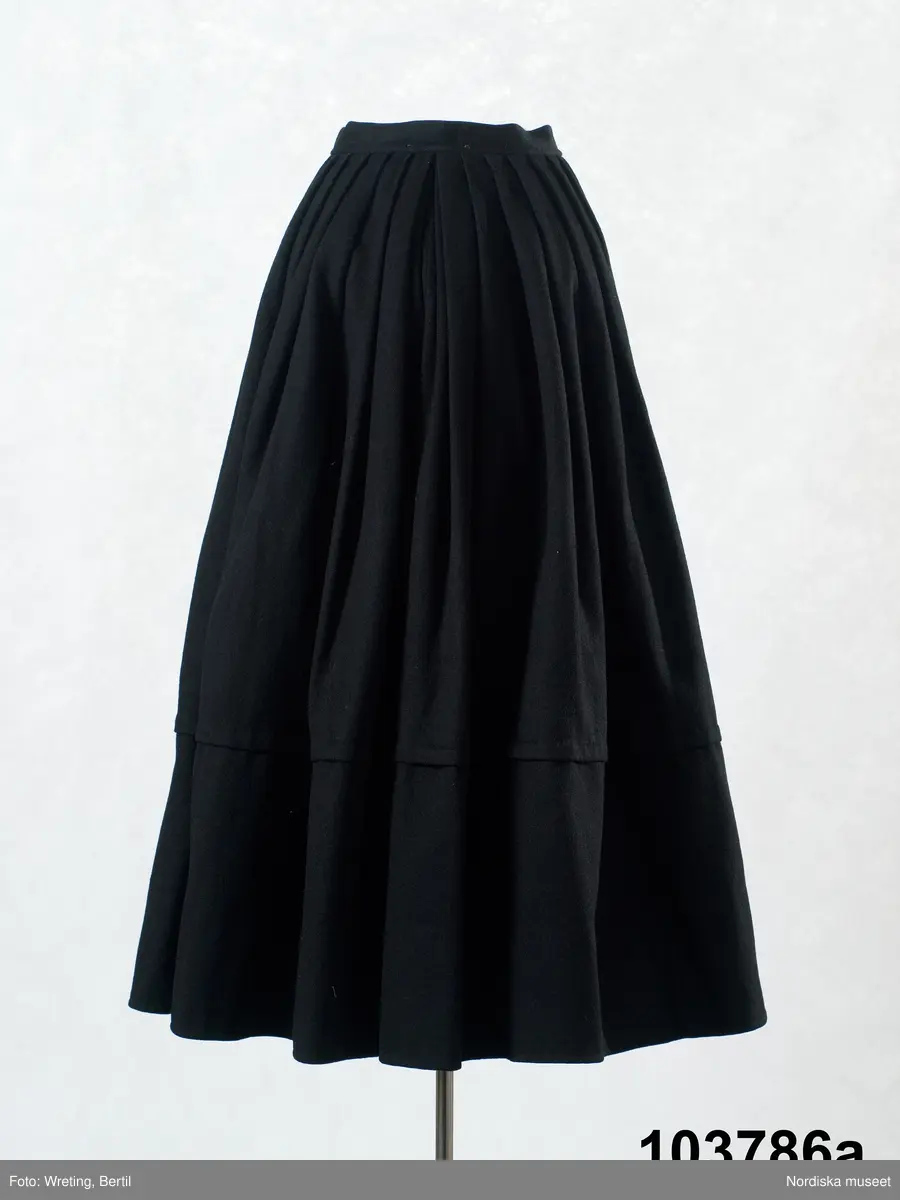 b. Brudtröja av svart vadmal,  Runt alla kanter bård av ytterst brunsvart sammetsband och innanför ett 4 cm brett svart sammetsband  med invävt bladmönster. På ärmarna ett svart slätt sammetsband med pålagd guldspets därefter siden band i rosa och grönt på botten av  svart  sammet och överst i bården en knypplad guldspets. 
c: Förkläde. Material: Textil, bomull. Teknik: Manufakturvävning. Handsömnad. Längd: 77 cm. Bredd: 110 cm. Färg: Vit.
Förkläde i vitt manufakturvävt bomullstyg, ihopsytt i mitten, smalrandigt, stärkt. Rynkningar mot midjelinning i tuskaftat bomullstyg, 1,5 cm bred, fastsydda bomullsband i sidorna. Uppvikt fåll nedtill, 6,5 cm, ovanför fåll, 5 lagda veck med ca 1 cm mellanrum, enkel utdragssöm i kanten av vecken.
/Inga-Lill Eliasson 2008-02-25

e: Krage. Längd: 47,5, cm - totallängd, bröststycke. Bredd: 33 cm - bröststycke. Höjd: 14 cm - kraghöjd, inkl spets. Material: Textil, bomull. Lingarn - spets. Bomullsgarn - broderi. Teknik: Tuskaft. Vitbroderi, Knyppling. Handsömnad.
Krage i tuskaftat vitt bomullstyg. Rektangulär duk, uppklippt halshål med fastsydd krage, 14 cm hög. Kragen har 3,5 cm bred fåll, enkel utdragssöm i kanten. Vitbroderi  i framkant,  plattsöm och kantsydda hål med infällt tyll, två sydda knapphål. Handknypplad spets fastsydd på långsida och korstsidor, 2,5 cm bred. Öppning fram, 24 cm långt sprund. Kragen är handsydd.
Lästips: Svensson Sigfrid, (1935). Skånes Folkdräkter, en dräkthistorisk undersökning 1500 - 1900. Stockholm: Nordiska Museets Handlingar: 3.
/Inga-Lill Eliasson 2008-02-19