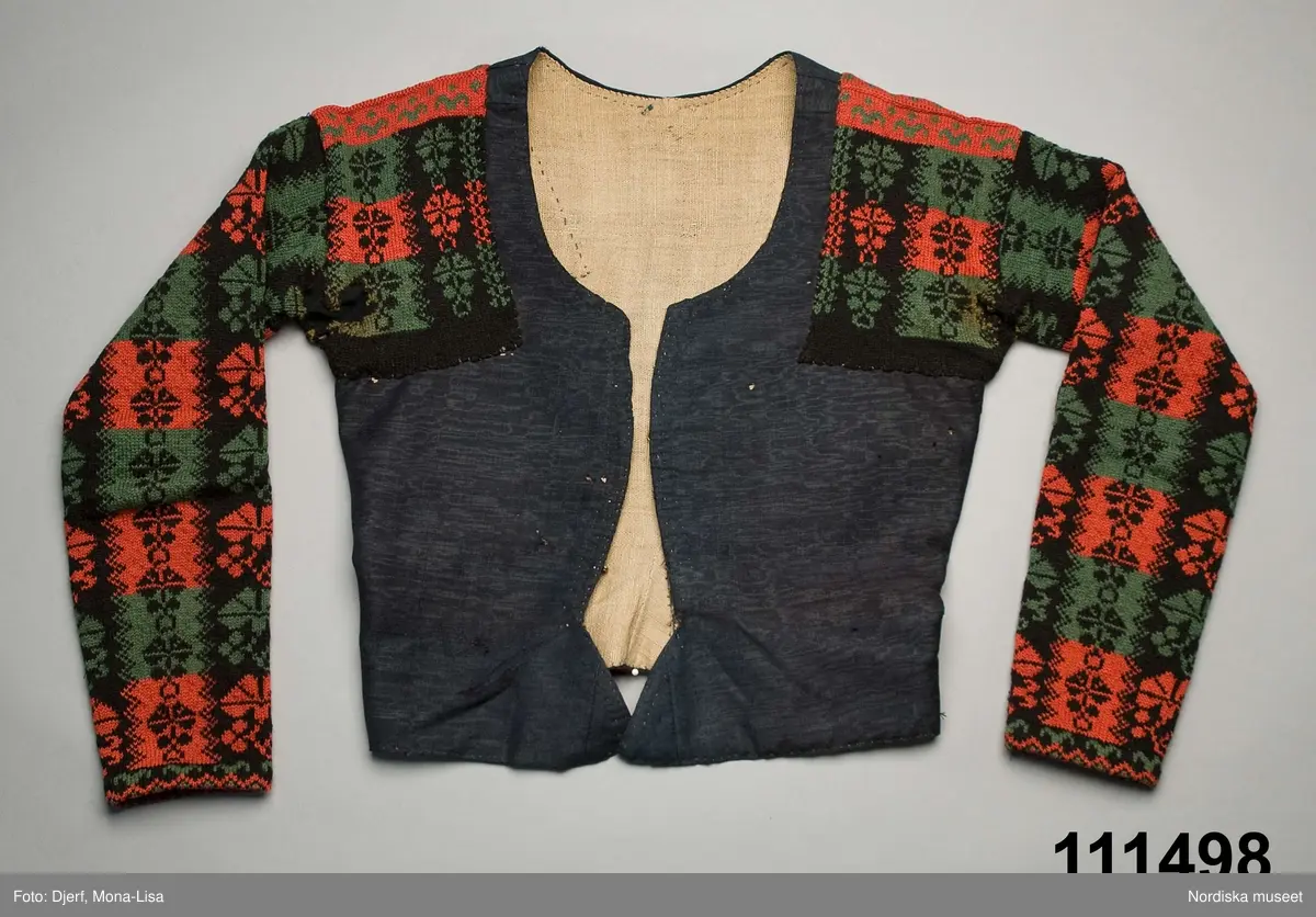 Ursprungligen ett livstycke av mörkblått kamgarnsylle i tuskaft med vattrad yta, s.k. kamlott. Livstycket i 1700-talssnitt med långt skört.
2 fram-och sidstycken, 2 bakstycken med 3 skörtveck i ryggen, 
Knäppning med 6 par hakar och hyskor, de av järntråd är äldst och ursprungliga, de i mässing yngre. Foder av grov oblekt linnelärft. Vid omsyning till tröja har skörtvecken sytts in i sömmarna ev. har hela livstycket även kortats något. Ärmarna mönsterstickade av rött tvinnat ullgarn och vitt bomullsgarn i stora rutor med nejlikmönster instickade i motsatsfärg. Ett rakt axelstycke stickat i samma mönstring och fastsytt vid ärmen och därefter påsytt på livstycket, så att det ursprungliga ärmhålet finns kvar under det stickade.
Anm. Ärmarna har fållats upp ca 2 cm

Kamlott var modetyg under senare delen av 1700-talet, det var ett dyrbart köpetyg och användes av allmogen i de finaste bröllopsplaggen som livstycken och kjol. Se t.ex svart kjol och tröja från Arbrå inv.nr 1824 och 1825. Ofta ser man sådana plagg omsydda till modernare snitt under 1800-talet. Ett så dyrbart och fint tyg återbrukades gärna. Jfr. liknande tröjor inv.nr 111497 och 201427 samt lösa ärmar 113011.
Troligen är samtliga dessa lösärmar stickade av samma person.
Beskrivning finns i boken Svenska Tröjor av Britt-Marie Christoffersson, ICA förlag 1988.
/Berit Eldvik 2006-06-16