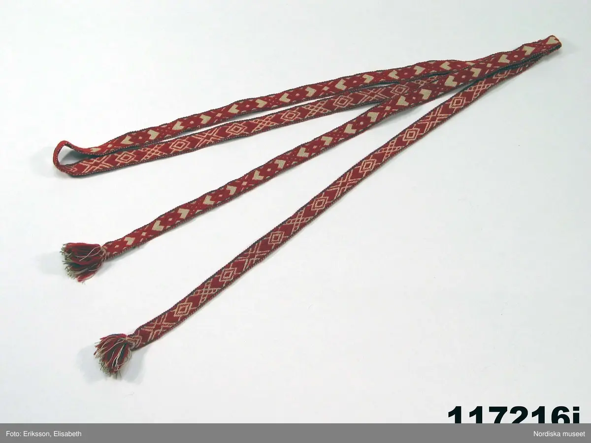 Huvudliggaren:
"Sorgdräkt för kvinna. Särk med 'hängseltåg', 'särkyvdel'; vit -'häsklä'; blått livstycke; svart tröja; huvudduk; 'svart näsduk'; hatt; 'raskförklä', 'sockar',  'hårbandsvifsa', huvudbindel."

Bilaga:
a: "8 Särk, övdel. av linne eller blågarn. med broderade linningar på överdelen"
c: "11. Hvit häsklä."
Anm: Kung Kerstin Larsdotters resa i Floda sn Dalarna 24/2 - 8/3 1912. Innehåller förteckning på inköp av dräktdelar från Floda, inv nr 117 216 - 117 239. Här finns  tillverkningstid och beskrivningar på vissa av dräktdelarna.  I vissa fall nämns också utförligt på vilka helgdagar dräktdelen  användes.

Katalogkort:
a: Hängselsärk
b. Överdel av halvlinne vävt i kypert.
c.  Halskäde av småmönstrat bomullstyg, liknande kambris, vävt  i kypertvariation med drällmönster.
d. Livstycke av mörkblå kraftig vadmal,
e. Tröja av grov svart vadmal

i. 
 Mönstervävt band med upplockat mönster i rött z-tvinnat ullgarn på vit linnebotten, med hjärtan, kryss och rombformer, 2 band om vardera 124 cm har vikts och sytts ihop så att det ena bildar en ögla och det andra har fria ändar avslutade med en tofs i bandets garn.
Läs mer om dräktskicket i Floda i: Albert Alm, Dräktalmanacka för Floda socken, Nordiska museet, Sthlm 1930
/Berit Eldvik 2005-04-14