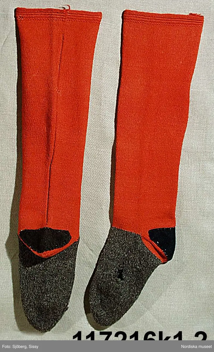 Huvudliggaren:
"Sorgdräkt för kvinna. Särk med 'hängseltåg', 'särkyvdel'; vit -'häsklä'; blått livstycke; svart tröja; huvudduk; 'svart näsduk'; hatt; 'raskförklä', 'sockar',  'hårbandsvifsa', huvudbindel."

Bilaga:
a: "8 Särk, övdel. av linne eller blågarn. med broderade linningar på överdelen"
c: "11. Hvit häsklä."
Anm: Kung Kerstin Larsdotters resa i Floda sn Dalarna 24/2 - 8/3 1912. Innehåller förteckning på inköp av dräktdelar från Floda, inv nr 117 216 - 117 239. Här finns  tillverkningstid och beskrivningar på vissa av dräktdelarna.  I vissa fall nämns också utförligt på vilka helgdagar dräktdelen  användes.

Katalogkort:
a: Hängselsärk
b. Överdel av halvlinne vävt i kypert.
c.  Halskäde av småmönstrat bomullstyg, liknande kambris, vävt  i kypertvariation med drällmönster.
d. Livstycke av mörkblå kraftig vadmal,
e. Tröja av grov svart vadmal

i. 
 Mönstervävt band med upplockat mönster i rött z-tvinnat ullgarn på vit linnebotten, med hjärtan, kryss och rombformer, 2 band om vardera 124 cm har vikts och sytts ihop så att det ena bildar en ögla och det andra har fria ändar avslutade med en tofs i bandets garn.
Läs mer om dräktskicket i Floda i: Albert Alm, Dräktalmanacka för Floda socken, Nordiska museet, Sthlm 1930
/Berit Eldvik 2005-04-14