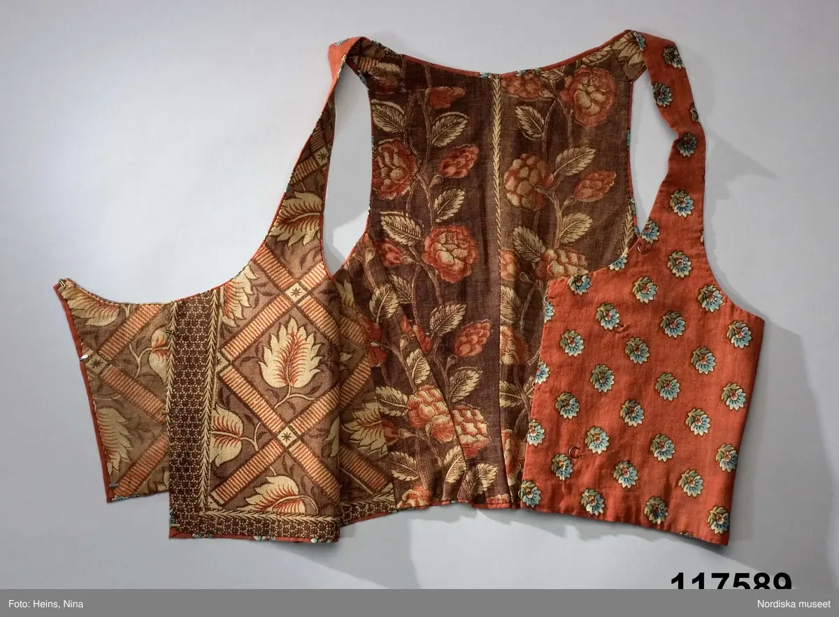 Livstycke av tryckt bomullstyg, kallat kattun, botten färgad i mörk krappröd färg med utsparade små blommor jämnt spridda över ytan. Blommorna har sedan tryckts i med blått och gult med svarta konturer. 2 fram/sidstycken och 4 ryggstycken som avsmalnar ned i ett rudimentärt skört utan veck, små infällda kilar i sidorna. Knäppt med 3 par hakar och hyskor, knäppningen dold under ett förslag av kattuntyget och en bit från ett halskläde i sidensatin med kantbården kvar. Livstycket fodrat med 2 olika kattuntryck av troligen något äldre datum, i ryggen ett brunbottnat bomullstyg med rosenrankor i krapprött och den oblekta grundfärgen, och i framstyckena ett bladmönster i gallerverk i samma färgskala men något ljusare. Troligen är det senare tyget också ett återbrukat halskläde där man skickligt lagt kantbården så att den följer även runt livstyckets kanter.
/Berit Eldvik 2006-12-08
