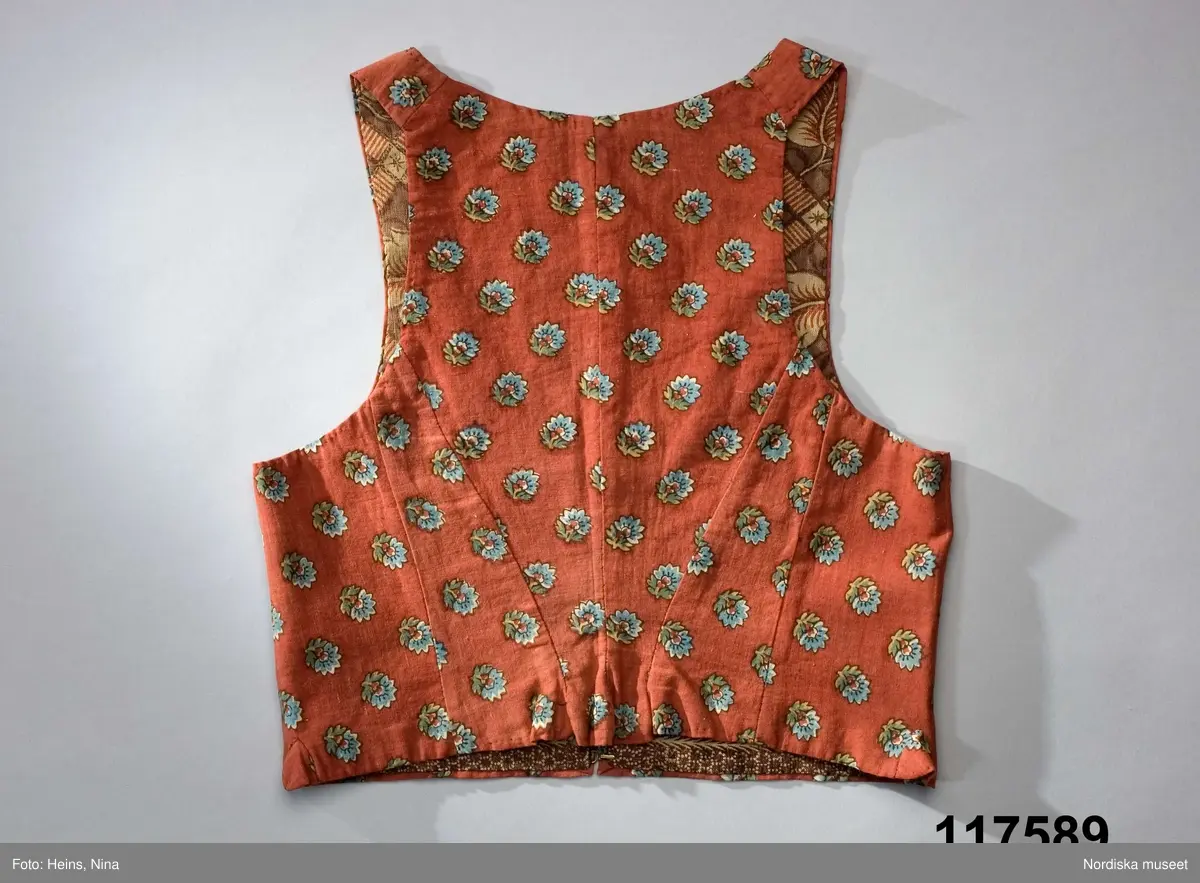 Livstycke av tryckt bomullstyg, kallat kattun, botten färgad i mörk krappröd färg med utsparade små blommor jämnt spridda över ytan. Blommorna har sedan tryckts i med blått och gult med svarta konturer. 2 fram/sidstycken och 4 ryggstycken som avsmalnar ned i ett rudimentärt skört utan veck, små infällda kilar i sidorna. Knäppt med 3 par hakar och hyskor, knäppningen dold under ett förslag av kattuntyget och en bit från ett halskläde i sidensatin med kantbården kvar. Livstycket fodrat med 2 olika kattuntryck av troligen något äldre datum, i ryggen ett brunbottnat bomullstyg med rosenrankor i krapprött och den oblekta grundfärgen, och i framstyckena ett bladmönster i gallerverk i samma färgskala men något ljusare. Troligen är det senare tyget också ett återbrukat halskläde där man skickligt lagt kantbården så att den följer även runt livstyckets kanter.
/Berit Eldvik 2006-12-08