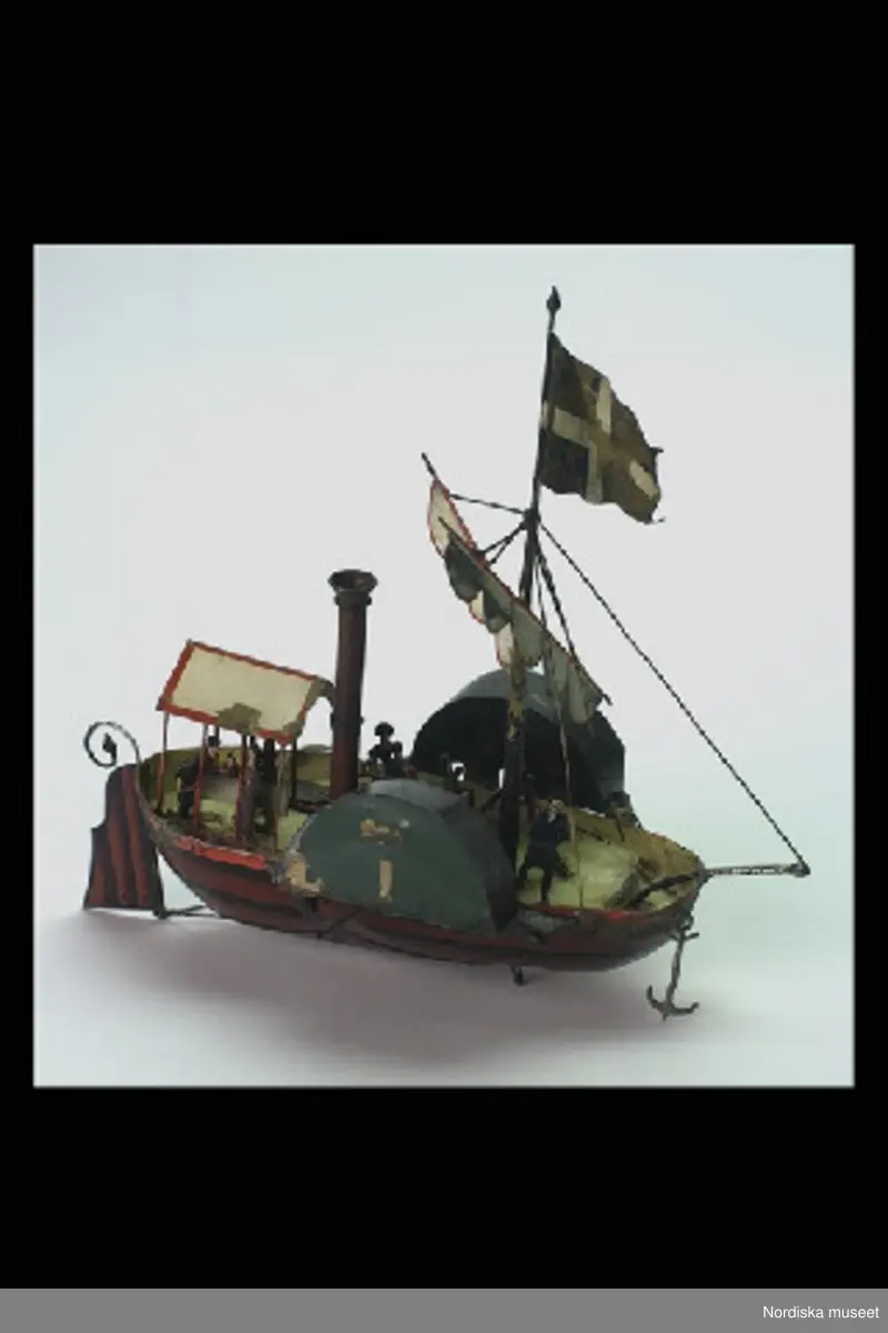 Katalogkort:
"Leksak, hjulångare med nyckel, mekanisk, bleckplåt, målad. 1800-talets mitt, Tyskland [med blyerts]. Spetsgattat fartyg, brunt skrov med svarta linjer; utanpåliggande roder, i fören ankare. gult däck; hög brun skorsten; mast med beslagna segel samt i toppen flagga. I aktern överbyggnad uppburen av tunnt rött galler. Vid friborden skovelhjul täckta av gröna och svarta skärmar. Däcket befolkat av 6 små figurer med målade dräkter  och anletsdrag. Urverksdriven, dras upp med liten fyrkantsnyckel av mässing; kvadratisk överdel med blomornament. /.../Tillstånd: fägen delvis avskavd. Masten avbruten. Förstagets infästning vid peket avbruten. AT 1982." [= Annika Tyrfelt ].
