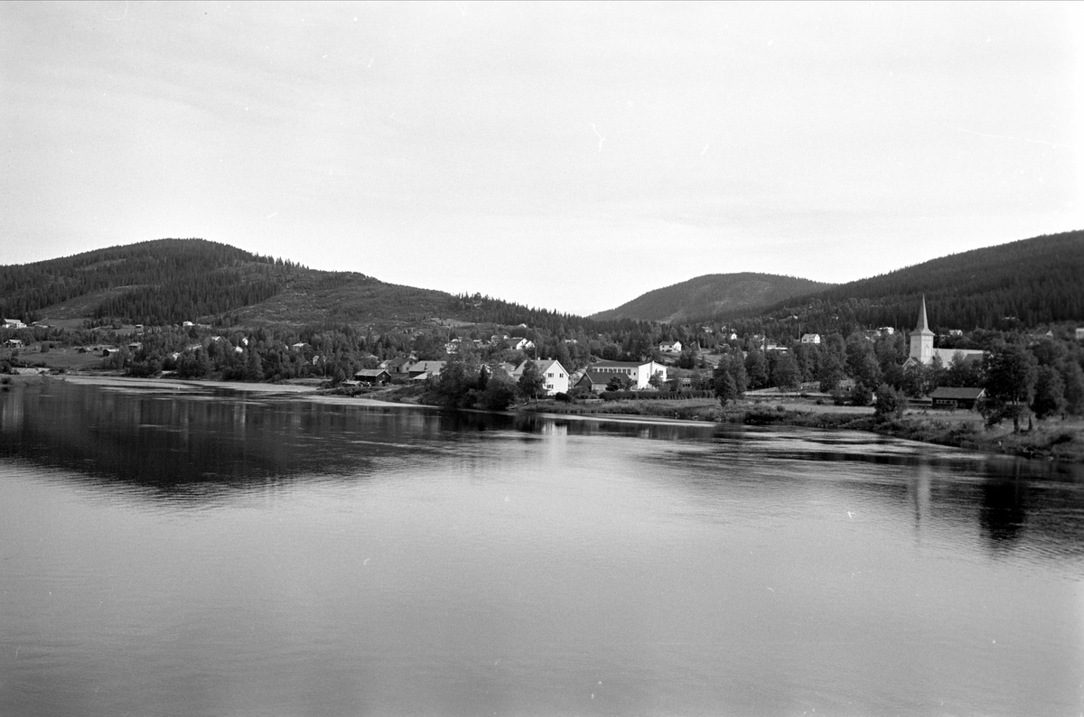 Trysil 1961. Utsiktsbilde med vann, Trysilelva, i forgrunnen. Innbygda med kirka i bakgrunnen.