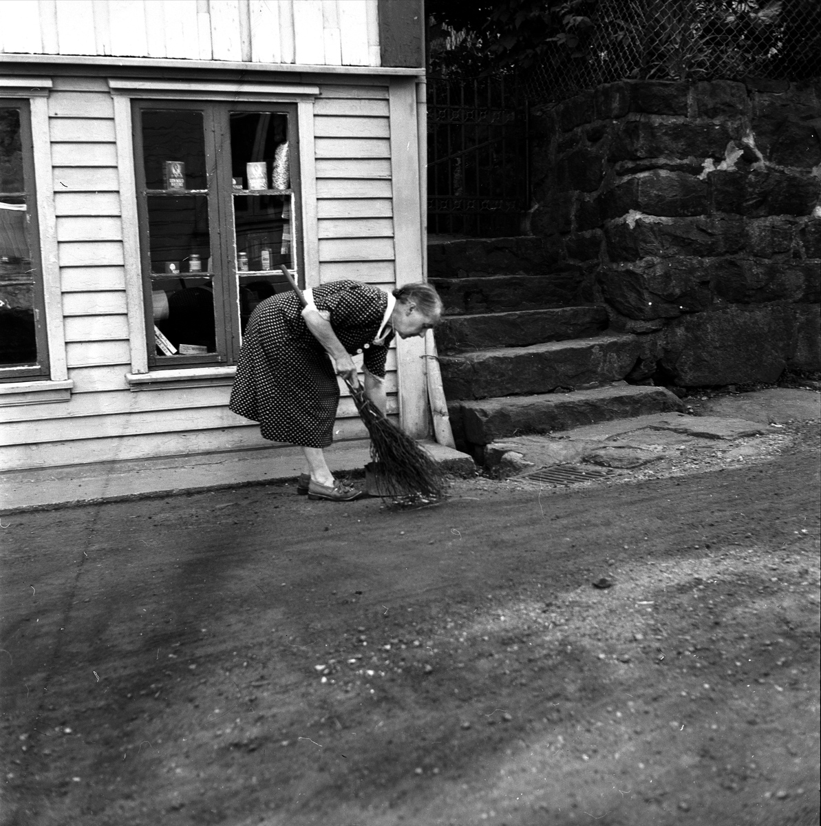 Sogndalsstranda, Sokndal, Rogaland, juli 1956. Kvinne med sopelime soper foran hus.