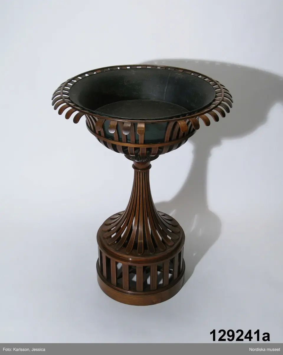 Huvudliggaren:
"blombord i from av en vas på fot, av mahogny, smala spjälor med inlagda ljusare ränder. Sign. 'Johan Axel Österlind Snickare i Stockholm'. Med lös sockel även av mahogny, stämplat under J under kunglig krona och plåtskål [lös].
Ink. 19/1 1917 Auktionshuset Hoving & Winborg, Stockholm. (auktion 11/12 1916). 410 kr."