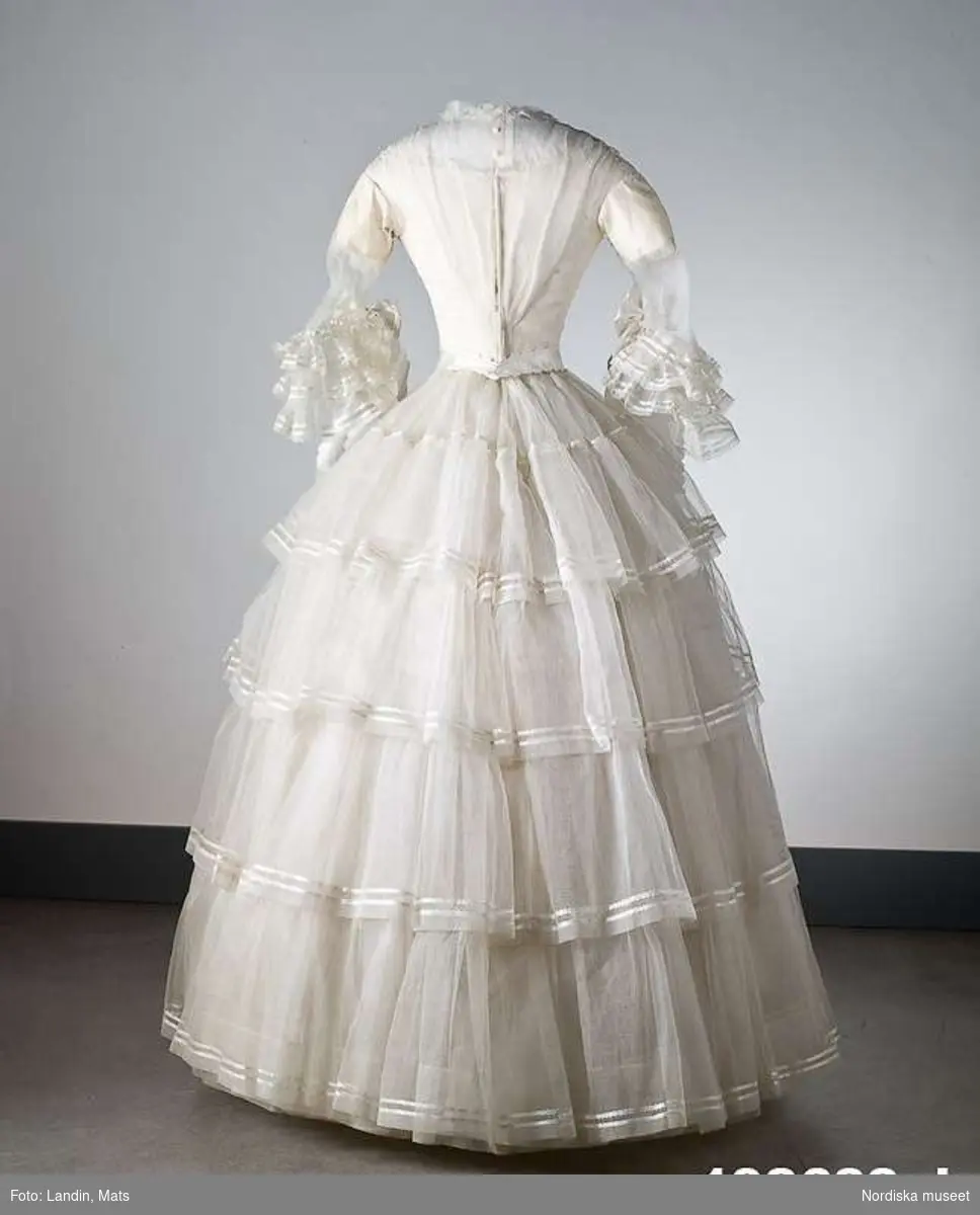 Brudklänning, liv med fastsydd kjol, vit tarlatan, garnerad med vita sidenband, rosetter. Klänningen har ett löst skärp av vit tarlatan.