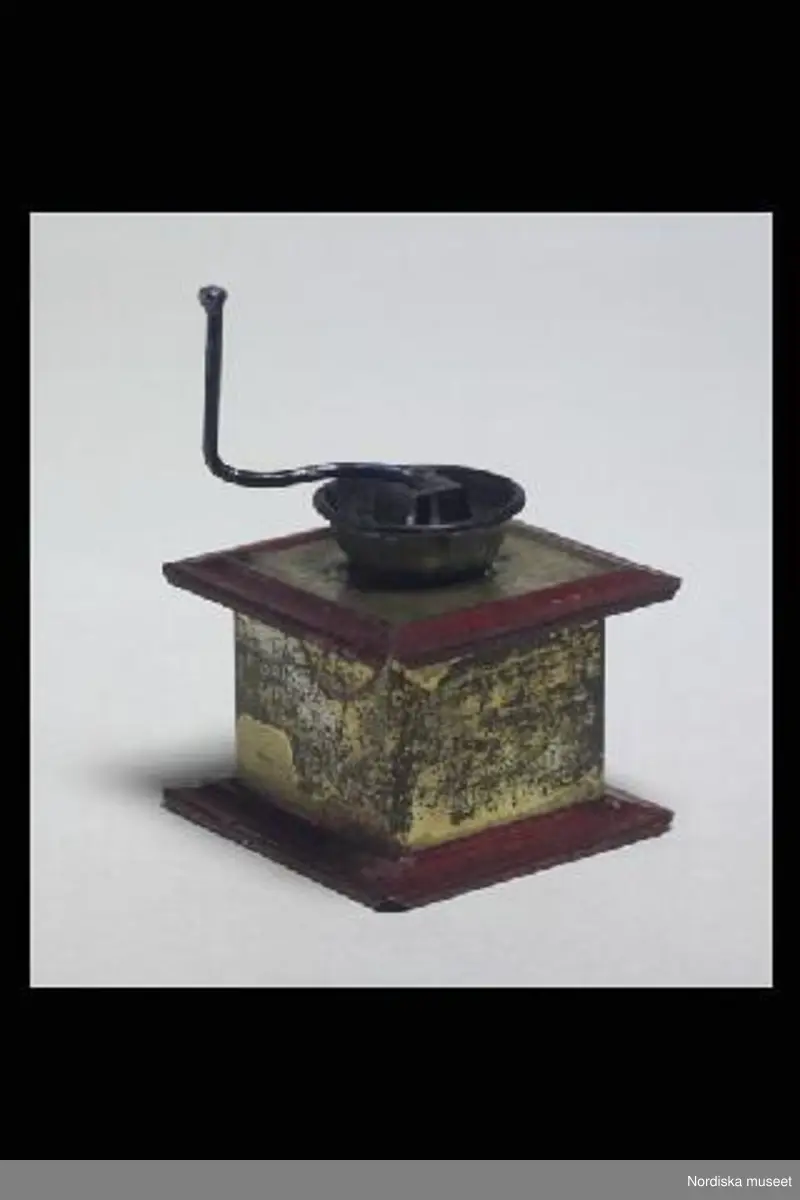 Inventering Sesam 1996-1999:
B 5   Dj 5   H 7,5  (cm)
Speldosa, kaffekvarn, leksak, av bleckplåt, kubiskt gullackerat hus, utskjutande rödlackerad bottenplatta och tak, tratt ooch rörlig vev av metall. 
Knäppande ljud när man rör veven.
Inköpt på auktion hos lanthandlare Lars Lundberg, Källeryd.
Birgitta Martinius 1997