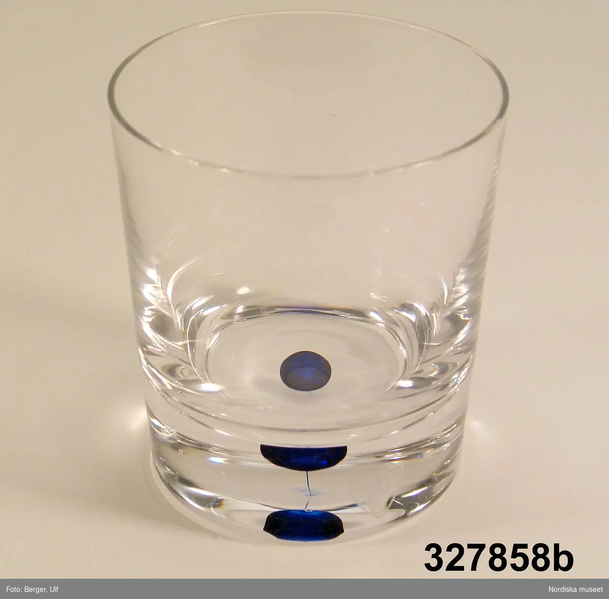 Huvudliggaren:
"Dricksglas i ask, 2 st, glas, papp. Klart, cylindriskt, koboltblå bubbla i tjock botten. Form Erik Lagerbielke Intermezzo, tillv 2000 Orrefors, etsad tillvsign. Vinröd ask, viklock. Folder. Anv 2001 under Sveriges EU-ordförandeskap. Old-Fashioned."

Kataloglapp:
Old fashioned-glas med tjock botten, koboltblå bubbla i botten.
Form Erika Lagerbielke, 1985 "Intermezzo"
Vinröd originalkartong.

Ingick i den kuvertuppsättning som utformades av Sveriges Lantbrukares Riksförbund (LRF) och användes vid ett antal av de officiella middagar som anordnades av svenska staten under Sveriges ordförandeskap i Europeiska unionen.. 
Se även inv nr

Ingår i ett större förvärv av utrustning och möbler som användes vid den officiella representationen i samband med Sveriges ordförandeskap i Europeiska unionen, våren 2001.
Se även inv nr

Se även Nordiska museets arkiv 6/2002 för informations- och dokumentationsmaterial.
/Helena Ulander/Cecilia Hammarlund-Larsson 2002
Förpackning: Munksjö förpackningar
/Heidi Henriksson/Kersti Wikström 2004
