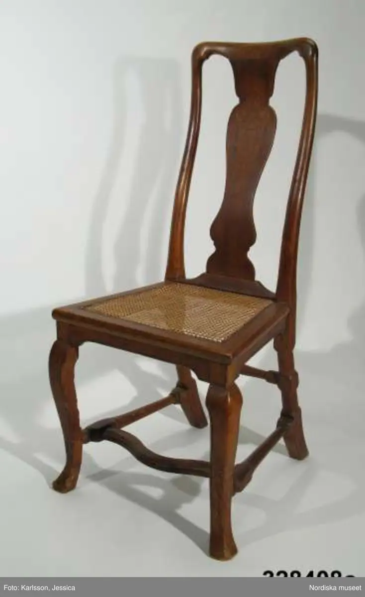 Stol, 6 st (a-f), England, 'Queen Anne', omkring 1720, av valnöt. Hög, balusterformad ryggbricka med inlagd (inkrusterad) dekor av ljusare träslag. Cabriolformade ben och svängt fotkryss med svarvade sidoslåar och bakre tvärslå. Sits med låg sarg, rottingflätad (rottingflätningen nygjord omkring 2002). Tillverkarinitialer inslagna på ben, ryggståndare och baksarg:
a) 'SH' (alt 'HS' ) samt 'IB'.
b) Rund stämpel: [kryss med dubbla streck] 
c) 'SH' (alt 'HS) samt 'IB'
d) 'SH' (alt HS') samt 'IB'
e) 'RH' (3 gånger) 
f) 'RH' (3 gånger),  'IB' (2 gånger) samt 'SH' (alt 'HS')

Jämför stol, inv nr 104.416 med initialerna 'RH' (enligt katalogkort). De engelska stolarnas märkning med tillverkarinitialer på t ex ben, sarg och ryggståndare visar på en rationalisering i tillverkningsprocessen. Initialerna är ibland placerade uppochnedvända vilket indikerar att de slogs in innan stolen sattes ihop. (se 'English furniture 1660-1714 - From Charles II to Queen Anne', Adam Bowett, Italien 2002, s. 264).
Anm: Flera stolar kompletterarade med delar av nytt trä, ilusade bitar. e) bakre tvärslå sekundärt förstärkt med skruv.
/Anna Womack 2008-09-25

Nordiska museet erbjöds 1992 att som testamentarisk gåva efter då framlidna Louise de Kiriline, Ontario, Kanada, ta emot en grupp sittmöbler bestående av två karmstolar och sex stolar. I samtal mellan arvinge till testator och dåvarande avdelningschefen Ingrid Bergman framgick att bakgrunden till gåvan var att del/delar av möblemanget redan skulle finnas i museets samlingar. En från Kanada översänd polaroidbild visade att en eller möjligtvis två stolar av denna typ, troligen tilllverkade i England omkr. 1720, förvärvats av museet redan 1905. Eftersom möbelgruppen skulle ge ett välkommet tillskott av engelska möbler till Charles Totties malmgård på Skansen tackade Nordiska museet ja till den erbjudna gåvan. Stolarna, som vid ankomsten till Sverige visade sig ha sekundära sitsar, har genom konservatorn Sören Lilja vid Skara museum åter försetts med rottingflätade sitsar av ursprunglig typ. Enligt testators anteckningar hade möblerna en gång tillhört den berömda romanförfattarinnan Sophie von Knorring (1797-1848). På sitt fäderne tillhörde hon ätten Zelow, som under 1700-talet haft stark förankring i Västergötland och även på hennes mans sida var det västgötska inslaget påfallande. De båda stolarna som redan tidigare tillhörde Nordiska museet, inv nr 104.415-104.416, inköptes 1905 från Göteborgs Historiska Museum dit de kommit från den kände samlaren Magister Nilsson i Skara. Dessa stolar är av samma typ, men olikheter i detaljer visar dock att de sannolikt ej ingått i samma möblemang. "Magister Nilsson", adjunkt vid Skara läroverk, som var en eldsjäl i Hazelius anda samlade kulturföremål från sin västgötska hembygd och de stora överensstämmelserna mellan dessa stolar och de stolar som nu förvärvas, visar att typen förekommit i Sverige under 1700-talet och kanske särskilt i de västra landskapen kring västerhavsporten Göteborg. Förvärvet behöver därför inte ses enbart relaterat till Tottieska malmgårdens möblering./Per Falck och Birgitta Martinius 2002.

Testator Louise de Kiriline Lawrence (född Flach, 1894-1992), var född i Sverige och växte upp på godset Svensksund på Vikbolandet i Östergötland. Efter att ha arbetat som röda korssyster under 1:a världskriget och i Ryssland efter revolutionen emigrerade hon till Kanada 1927. 