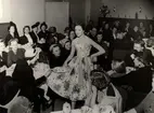 Visning Italiana på NK (Nordiska Kompaniet) 1953. Kvinnlig modell iklädd ljus klänning med bara axlar och blommönster. Modellen står i rum med publik som sitter vi små bord dukade med kaffe- eller tekoppar och kannor.