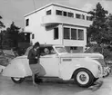 Kvinna i samtal med en passagerare i en vit Plymouth 1939, cabriolet, parkerad framför ett hus i funktionalsitisk stil. Nordiska Kompaniet.