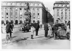 Stadsbild från Gustav Adolfs torg i Stockholm, 1910. Mitt i rondellen står en polis som övervakar trafiken, några kostymklädda herrar och en pojke i sjämanskostym med korta byxor. Cyklister, en bil och hästdragna kärror samsas på gatan. I bakgrunden byggnader och L'Archevêques staty förställande Gustav II Adolf.