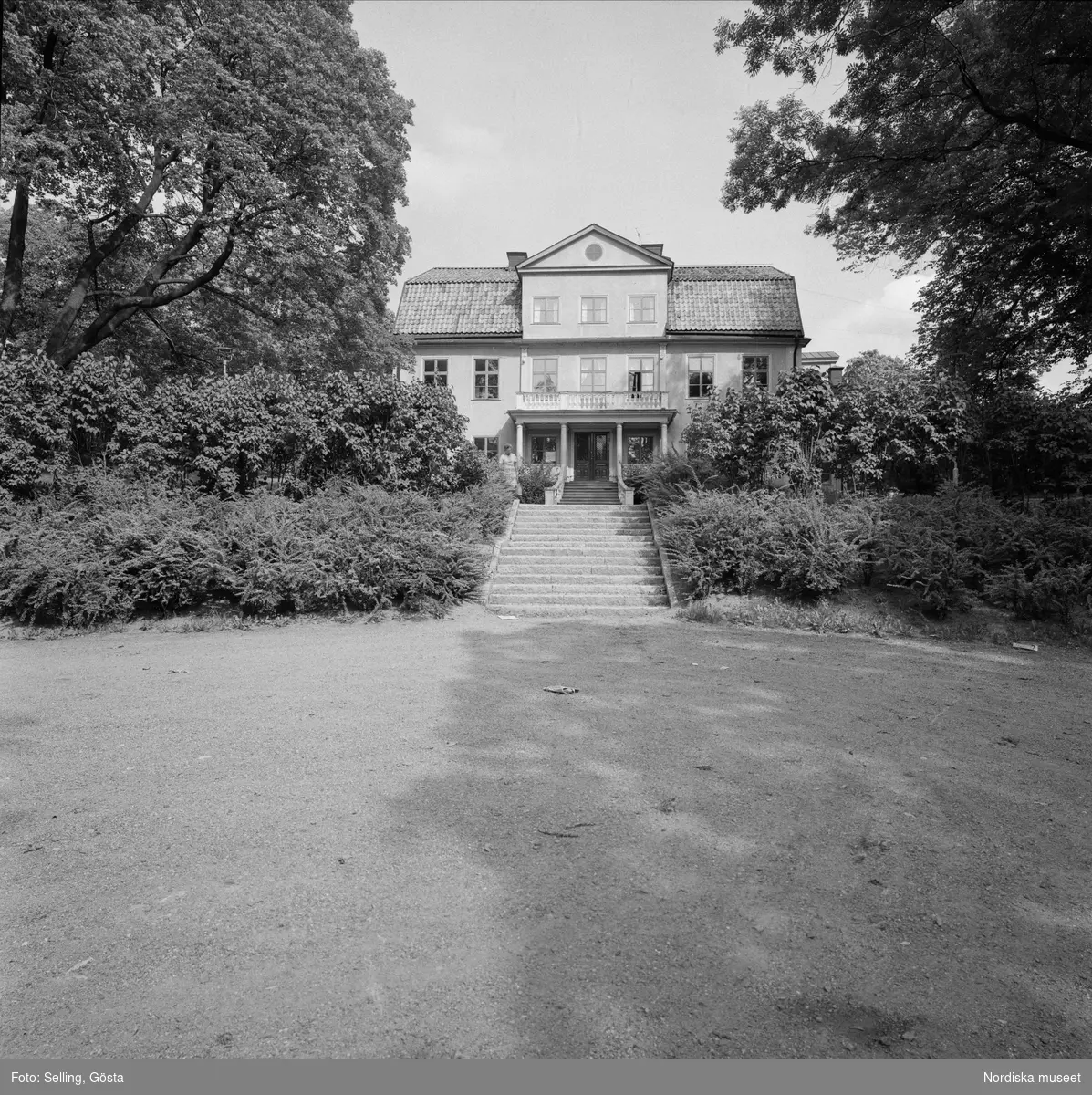 Enskede, gårdsfasaden från väster. Vid trappan står en kvinna bland grönskande buskar. I förgrunden parken. Brännkyrka socken, Svartlösa härad, Södermanland. 

