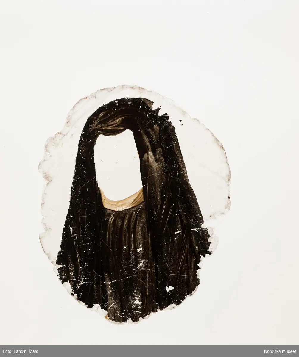 Katalogkort:
"Målning på kopparplåt, osign. Enligt uppgift av givaren föreställande en fröken Hahne. I fodral av svart pressat läder på stomme av papp med 13 glimmerskivor med målade dräkter och dräktdetaljer enligt uppg. målade av prof. N. H. Sjöborg (1767-1838)."
"Blont hår, mörkgrå ögon, rosafärgad klädning besatt med vita spetsar och spetskrage, vitt pärlband i hår och om hals, dessutom juvelhalsband och juvelbrosch. Bakgrund t.h. brunt draperi (vägg?) t.v. träd mot molnig blå himmel, 2 flygande fåglar. Kopparplåten genomborrad upptill. Till detta höra 13 st glimmerskivor att lägga på målningen. I svart ovalt etui, 88 mm x 73 mm av svart refflat skinn, invändigt klätt med ljusblått siden."

Genom att lägga de olika målade glimmerskivorna ovanpå porträttet förändras Kerstin Hahnes utseende. Skivorna visar enligt katalogkort:
- vit turban virad med gulbrunt, ljusbrun draperad dräkt med svarta prickar
- svart huvudbonad och gula örhängen
- svart huvudbonad, ljusröda örhängen, ljusröd bröstrosett
- svart hatt, svart kort jacka, vit krage och vita ärmar synliga i uppslitsningarna
- svart dok och klädning, vit krage
- ljusröd hårprydnad, örhängen vita och ljusröda, svart klädning med vit krage
- ljusbrunt hårnät, dito örhängen med vita pärlor, ljusbrun klädning med vita ärmar
- vitt nunnedok, svart dräkt
- ljusbrun hårprydnad, möjligen nät av band, vita örhängen, svart klädning, vit krage
- brun huva med krage, möjligen kappa
- svart barett med en grön, en vit och en rosafärgad plym, fästade med en gul agraff, vita örhängen, vit bröstrosett
- hårklädsel av 3 gula fjädrar samt en gul, en vit, och en grön plym, svart mask, vita örhängen, vitt band som täcker hakan, två gula bröstrosetter
- hårprydnad (nät?) ljusrött, röd och vita örhängen, till venster en neger, som omfamnar damen, under negerns huvud något gulvitt, som möjligen tillhör hans dräkt

Kerstin Hahne, död 1656, gift med Honorat Verdelet, var dotter till uppvaktaren hos kung Karl IX:s barn, Christoffer Hahne till Hesselby och Ingrid Björnsdotter.