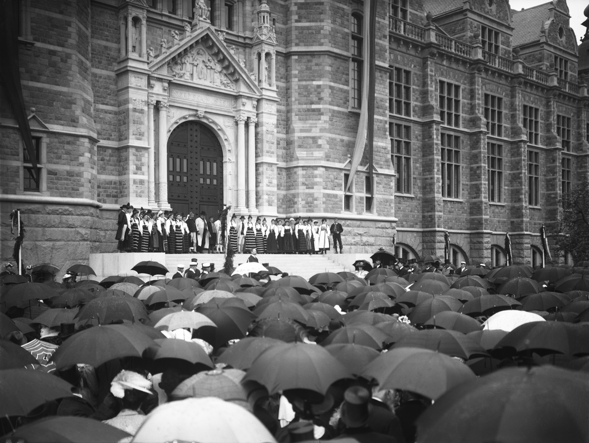 Nordiska museets invigning 1907. Museets medarbetare står överst på byggnadens trappa iförda folkdräkter. Byggnadens arkitekt, professor Isak Gustaf Clason står nedanför översta trappan och håller tal till åskådarna. Prinsessan Ingeborg, kronprins Gustav och prins Gustav Adolf skymtas bland åskådarna till höger om trappan. Åskådare med paraplyer i förgrunden. 