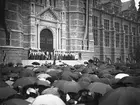 Nordiska museets invigning 1907. Museets medarbetare står överst på byggnadens trappa iförda folkdräkter. Byggnadens arkitekt, professor Isak Gustaf Clason står nedanför översta trappan och håller tal till åskådarna. Prinsessan Ingeborg, kronprins Gustav och prins Gustav Adolf skymtas bland åskådarna till höger om trappan. Åskådare med paraplyer i förgrunden.