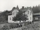 Falerum, Gärdserum sn, Småland. En kvinna med tre barn och en man bredvid två kvinnor framför en fastighet med ekonomibyggnad i bakgrunden.