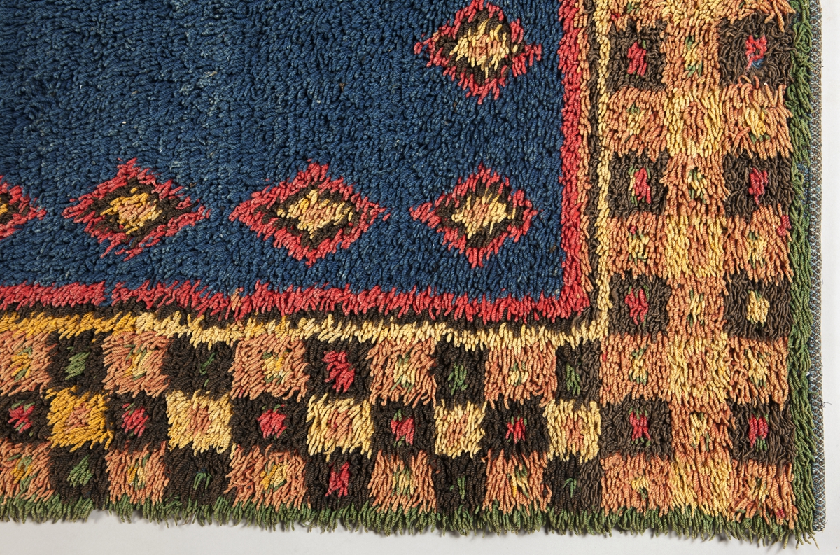 Prydnadsrya i två våder, hopsydda kant i kant. Botten i korndräll med varp av blått s-tvinnat lingarn 11 tr/cm och inslag av mörkbrun s-spunnen ull 7½ inslag/cm. Nockor av en tråd tvåtrådigt z-tvinnat hårdtvinnat ullgarn, knutna med giordesknut över 4 varptrådar med 4 varptrådar mellan nockorna och 4 inslag mellan nockraderna. Nockorna syns ej på baksidan.

Mönster: Stort blått mittfält avgränsat mot bården av en röd, en grönbrun och en gul smal linje. På det blå mittfältet, utmed sidorna romber av rött, grönbrunt och gult. Bred bård av schackrutor i grönbrunt, gult och rosagult. Gröna, röda och gula prickar i rutorna. Ytterst smal grön rand.